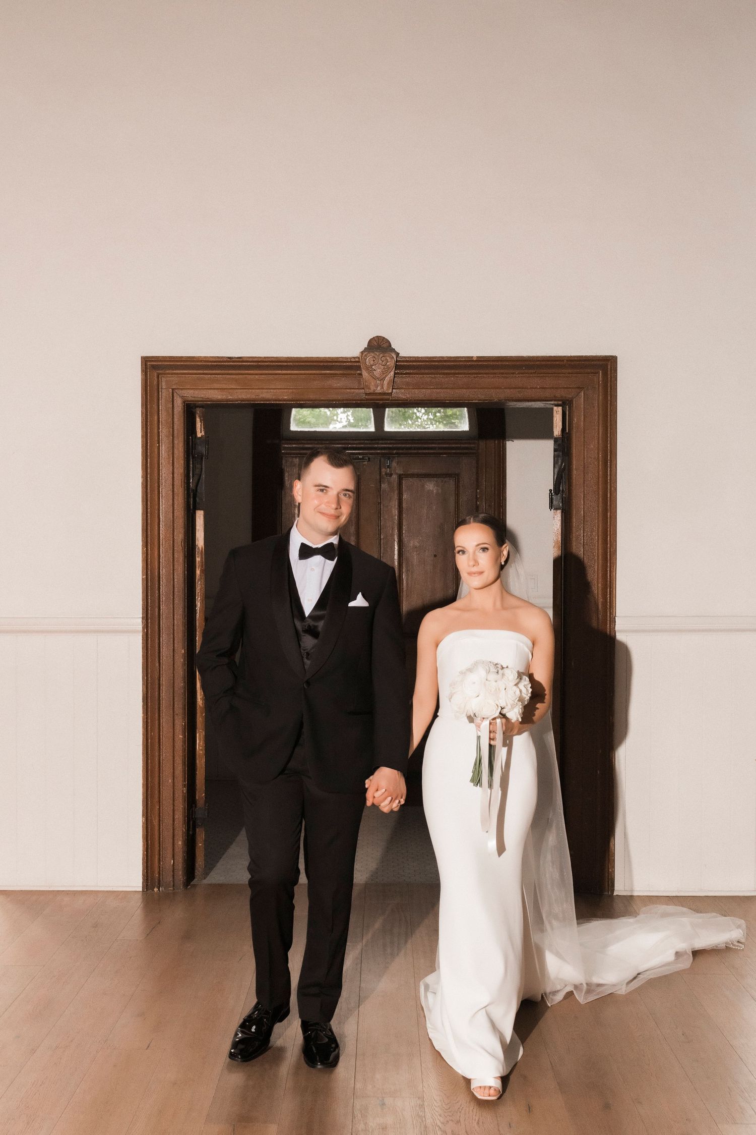 A couple walks through a wooden doorway wearing formal wedding attire in an elegant indoor setting.