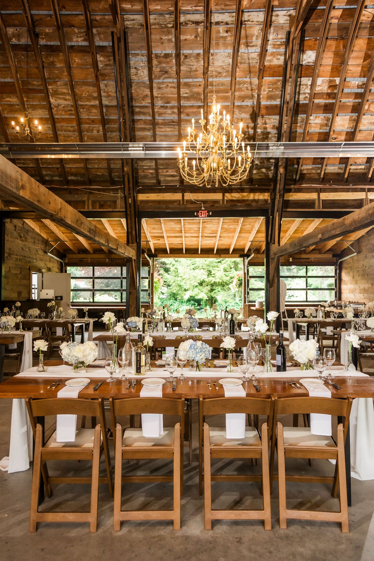 Rustic wedding reception venue with long wooden tables, elegant chandelier, and exposed beam ceiling in industrial-chic setting.