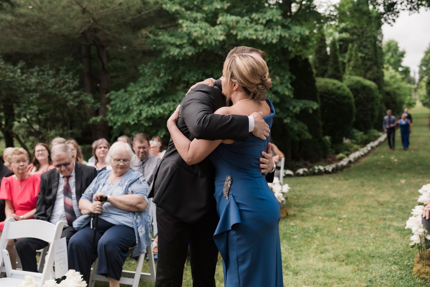 Two people embrace in a warm hug at an outdoor wedding ceremony while guests look on from white folding chairs.