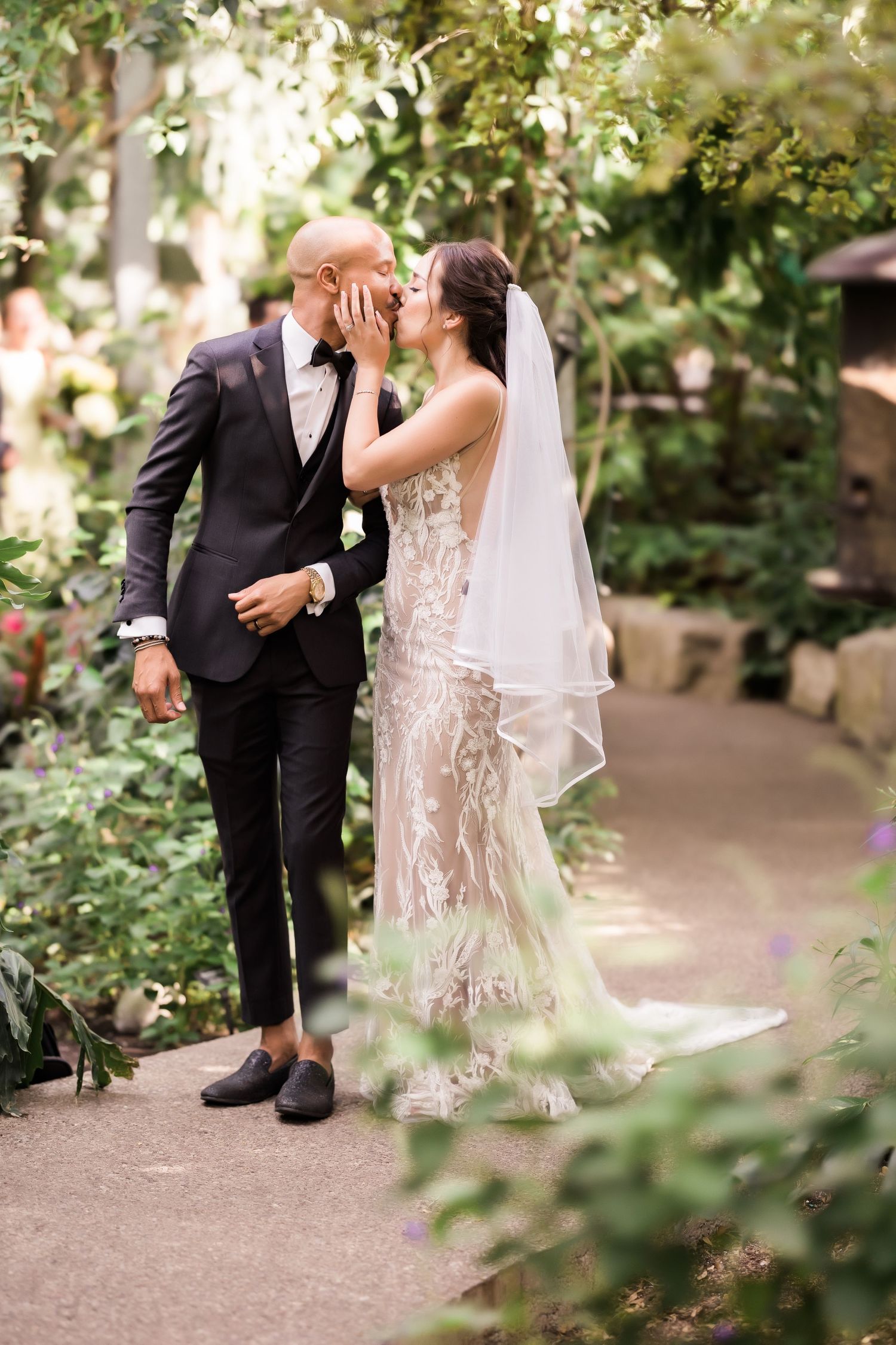 Newlywed couple shares intimate moment on garden path with bride in lace gown and groom in dark suit.