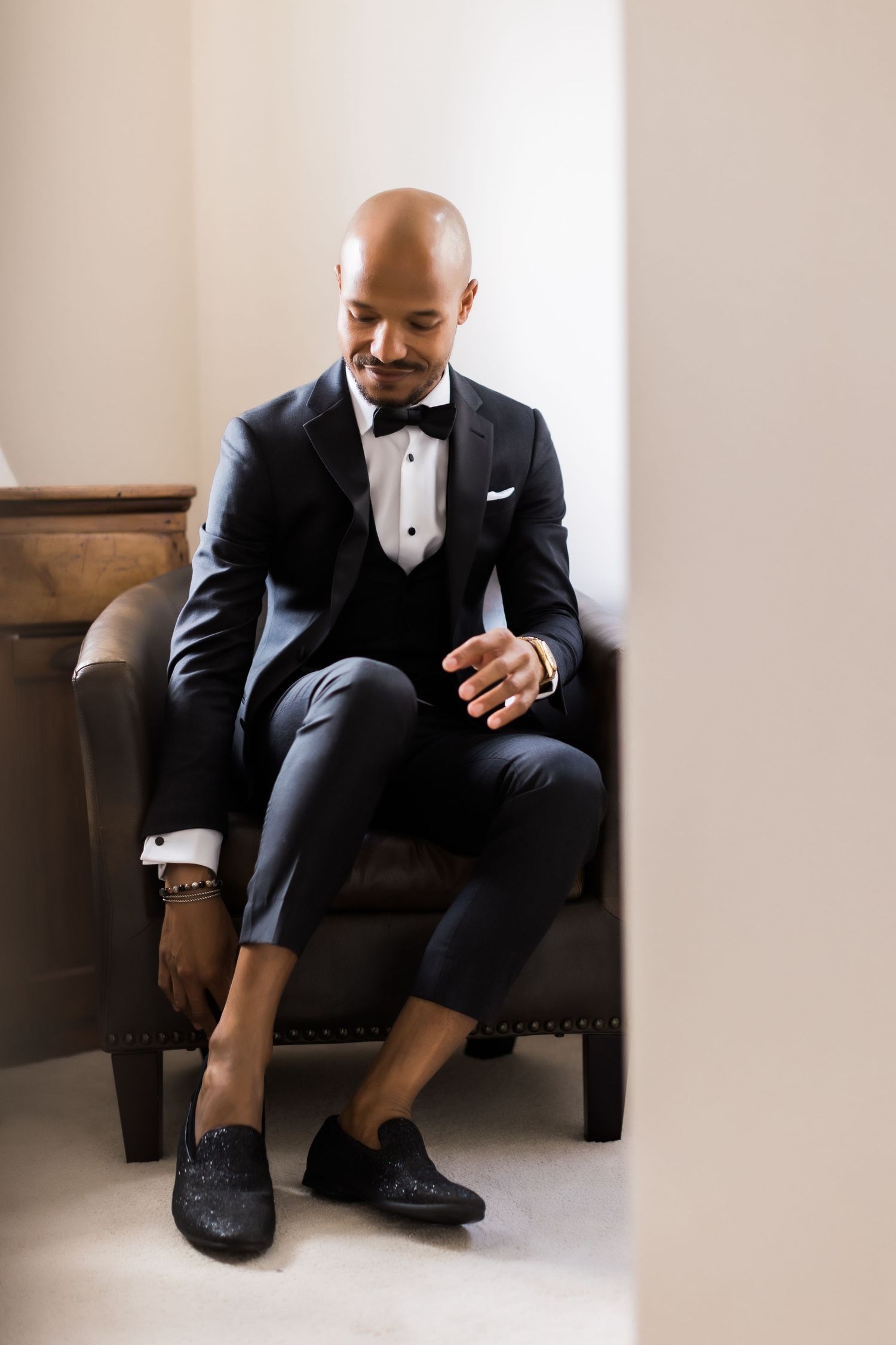 Elegant groom in black tuxedo and bow tie sits thoughtfully in brown leather chair.