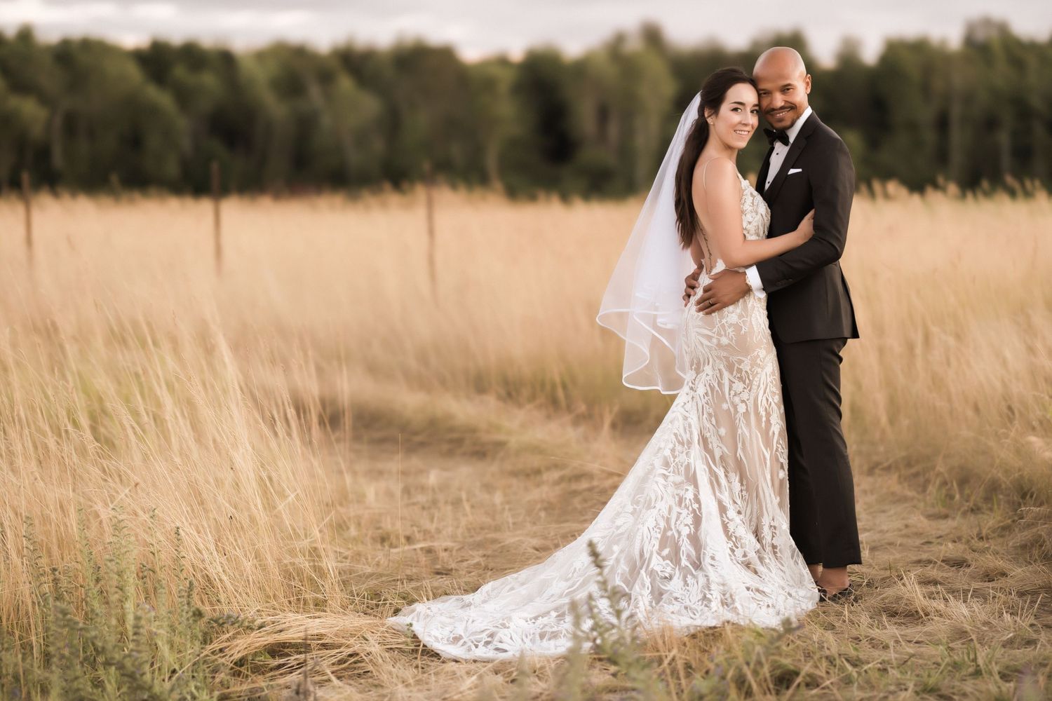 Wedding couple poses intimately in a sun-lit meadow with tall grass, bride wearing an elegant lace dress.