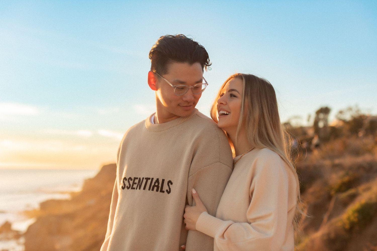 Happy couple in cream sweaters share laugh together on scenic coastal overlook.