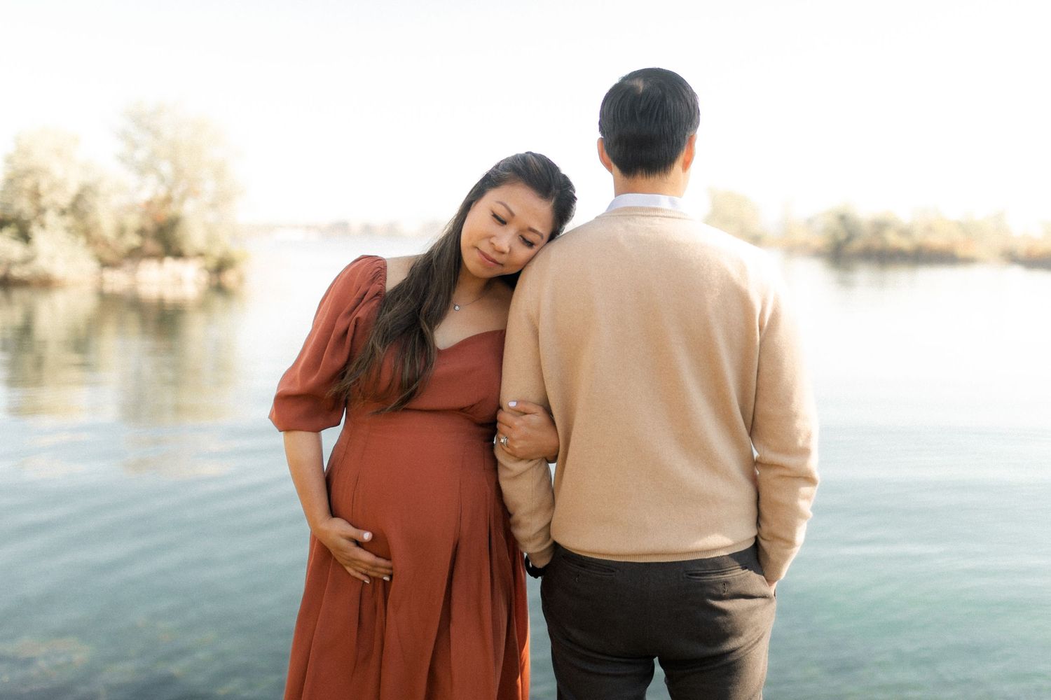 Tender maternity moment captured as couple stands by scenic lake.