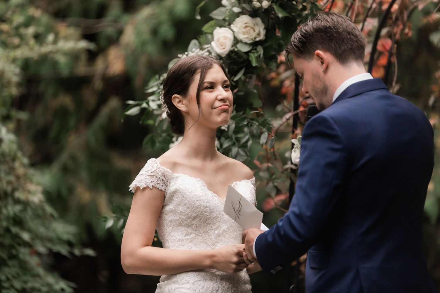 Wedding ceremony under floral arch with officiant leading service in natural outdoor setting.