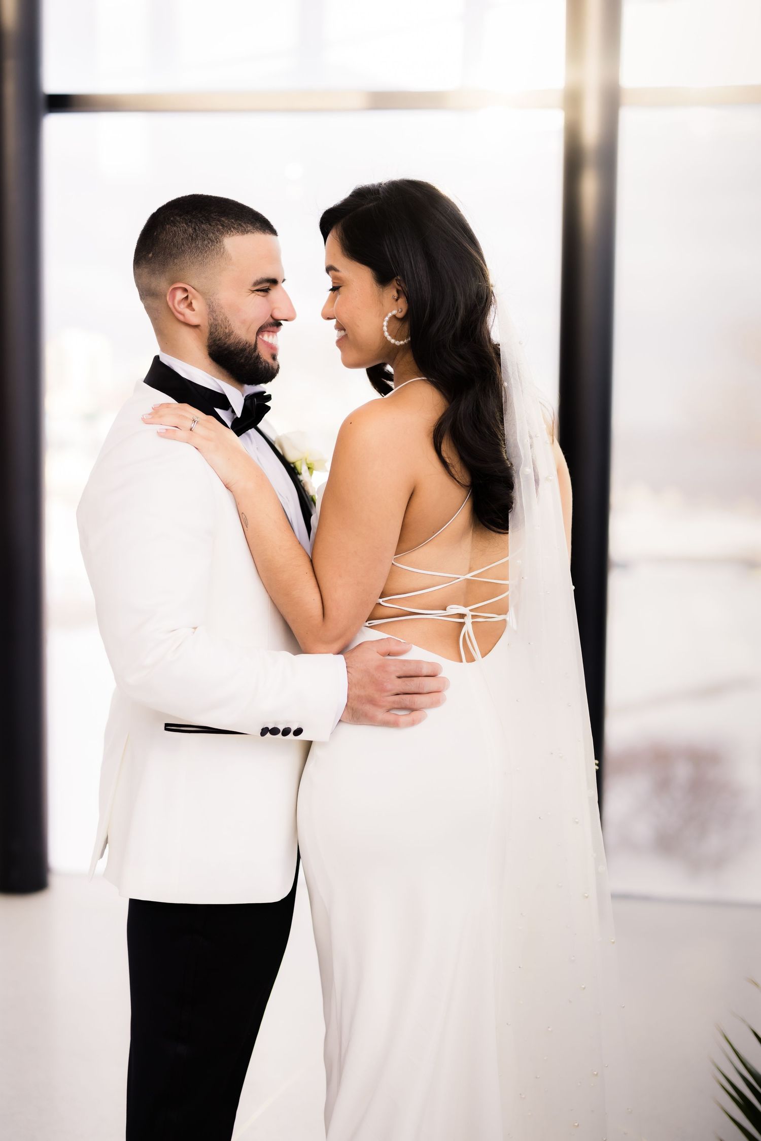 A romantic wedding portrait of a couple in white embracing each other near a bright window.