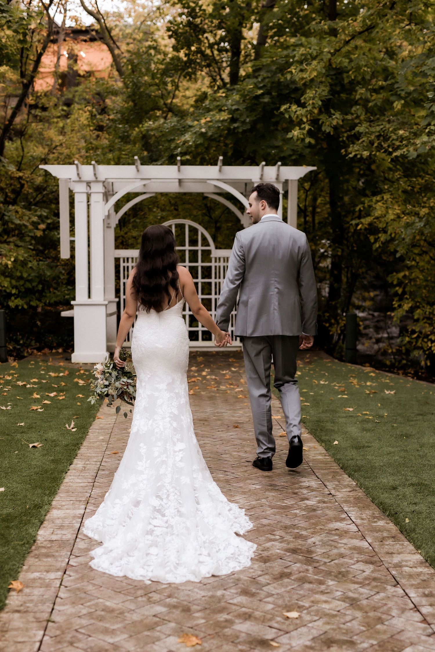 Couple walks toward white garden arbor on stone path surrounded by autumn foliage.