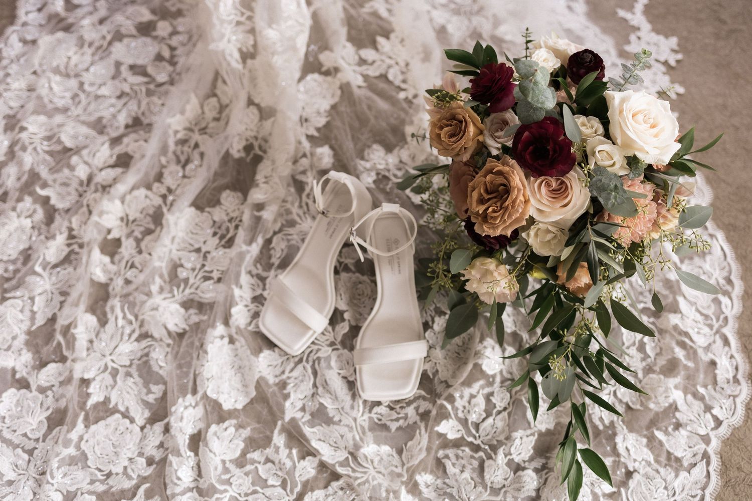 White bridal shoes and a romantic bouquet of blush and burgundy flowers placed on lace fabric.