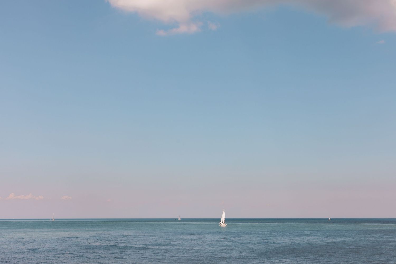 A minimalist seascape with a single white sailboat on blue waters under pastel skies.