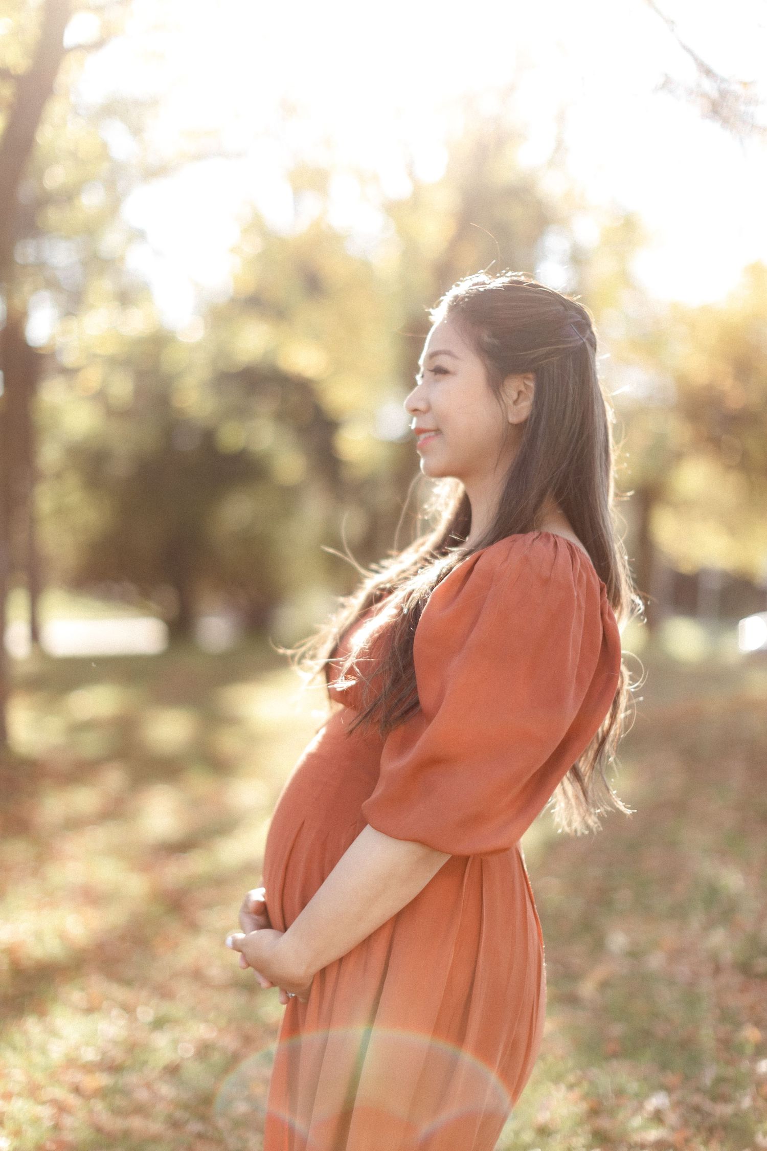 Expectant mother in rust-colored dress poses in profile against sunlit autumn trees.