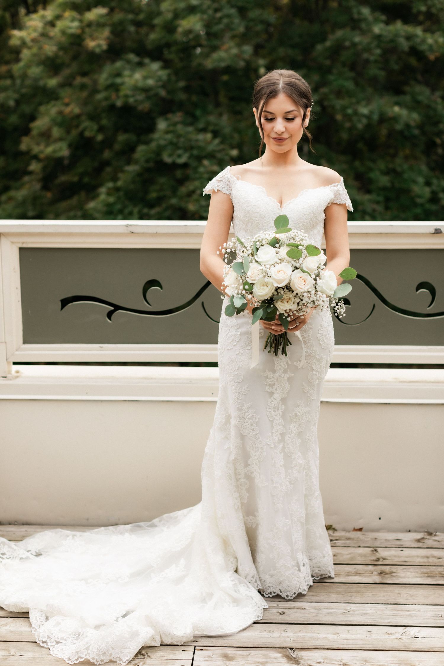 Elegant bride in lace gown holds bouquet on wooden deck with ornate railing.