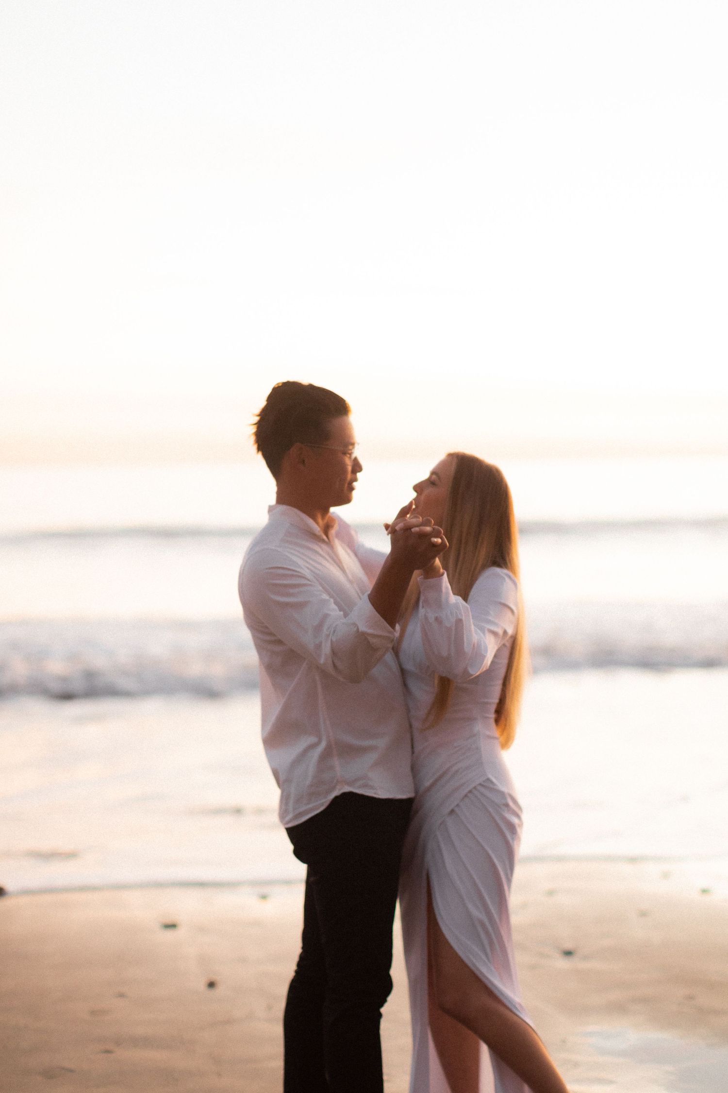 Couple in white outfits dancing together on beach during golden hour photography session.