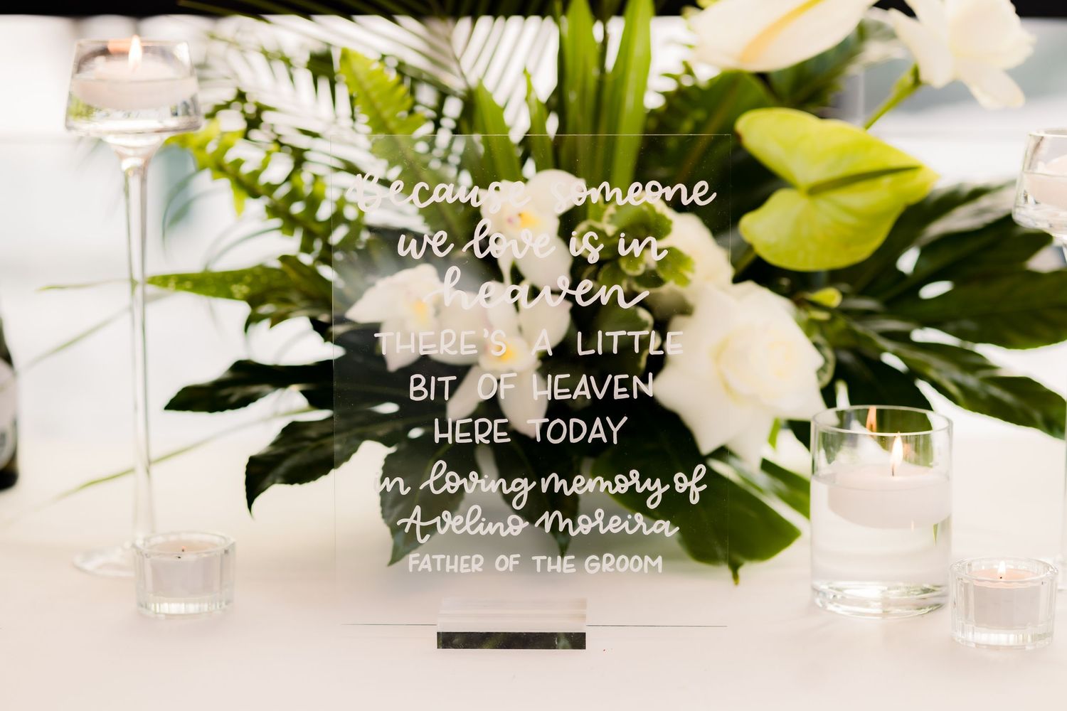 Clear acrylic wedding sign with white script text displayed on white tablecloth.