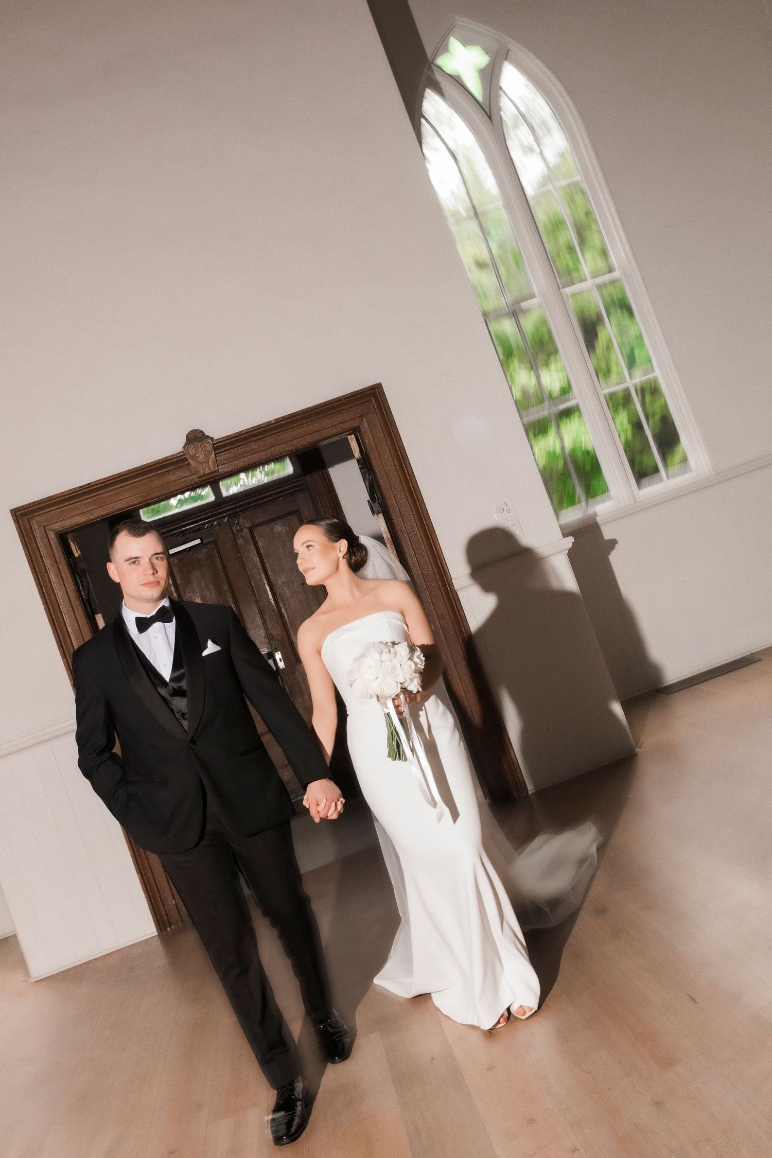 A couple in formal wedding attire pose near an arched window in a light-filled interior space.