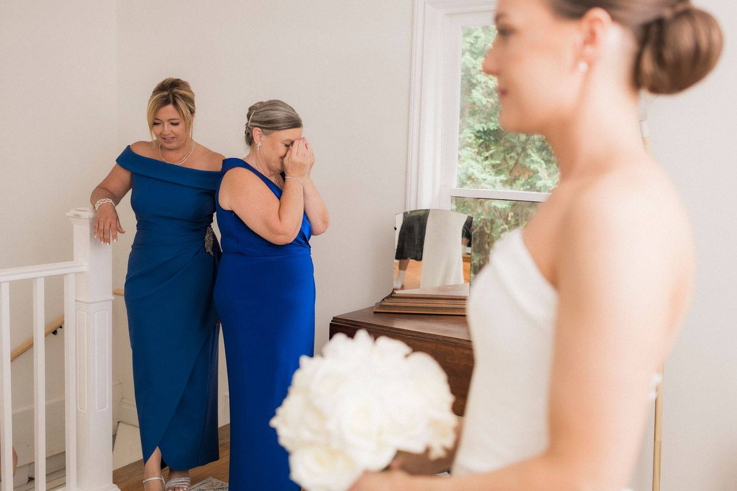 Two bridesmaids in royal blue dresses show emotion as they see bride in white gown holding white bouquet.