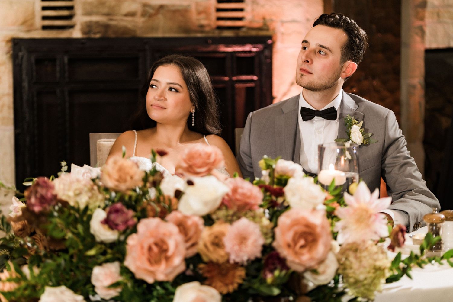 Wedding couple listens intently during reception with elegant peach rose centerpiece in foreground.