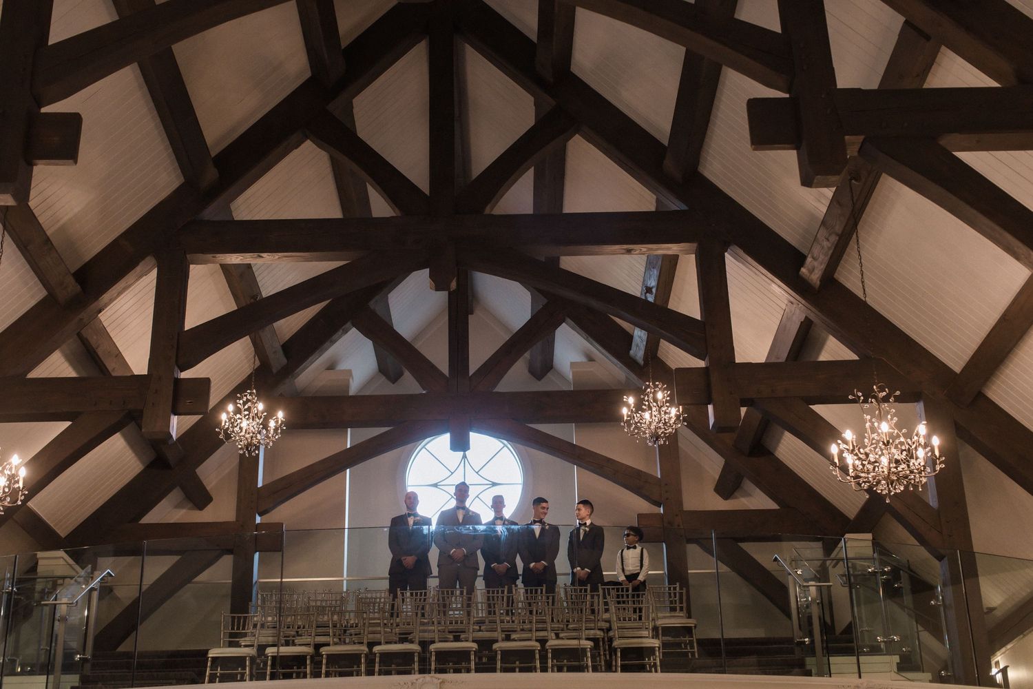 Groomsmen stand silhouetted against a circular window in a chapel with exposed wooden beams and crystal chandeliers.
