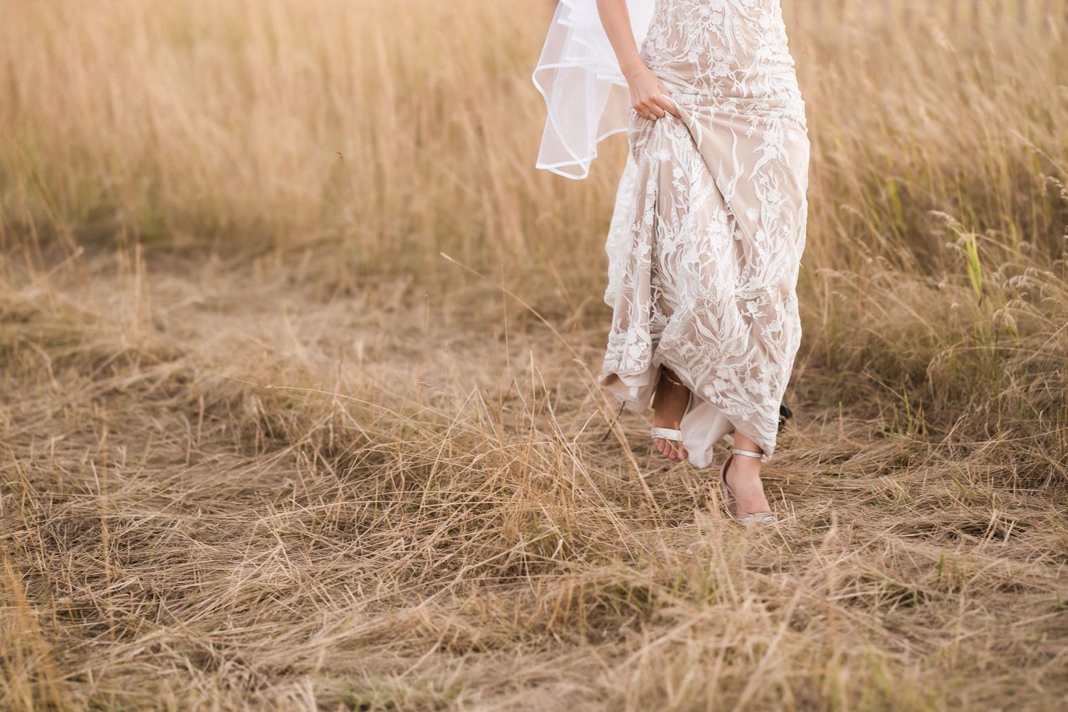 Close-up of bride's lace dress and bare feet walking through tall grass in field.
