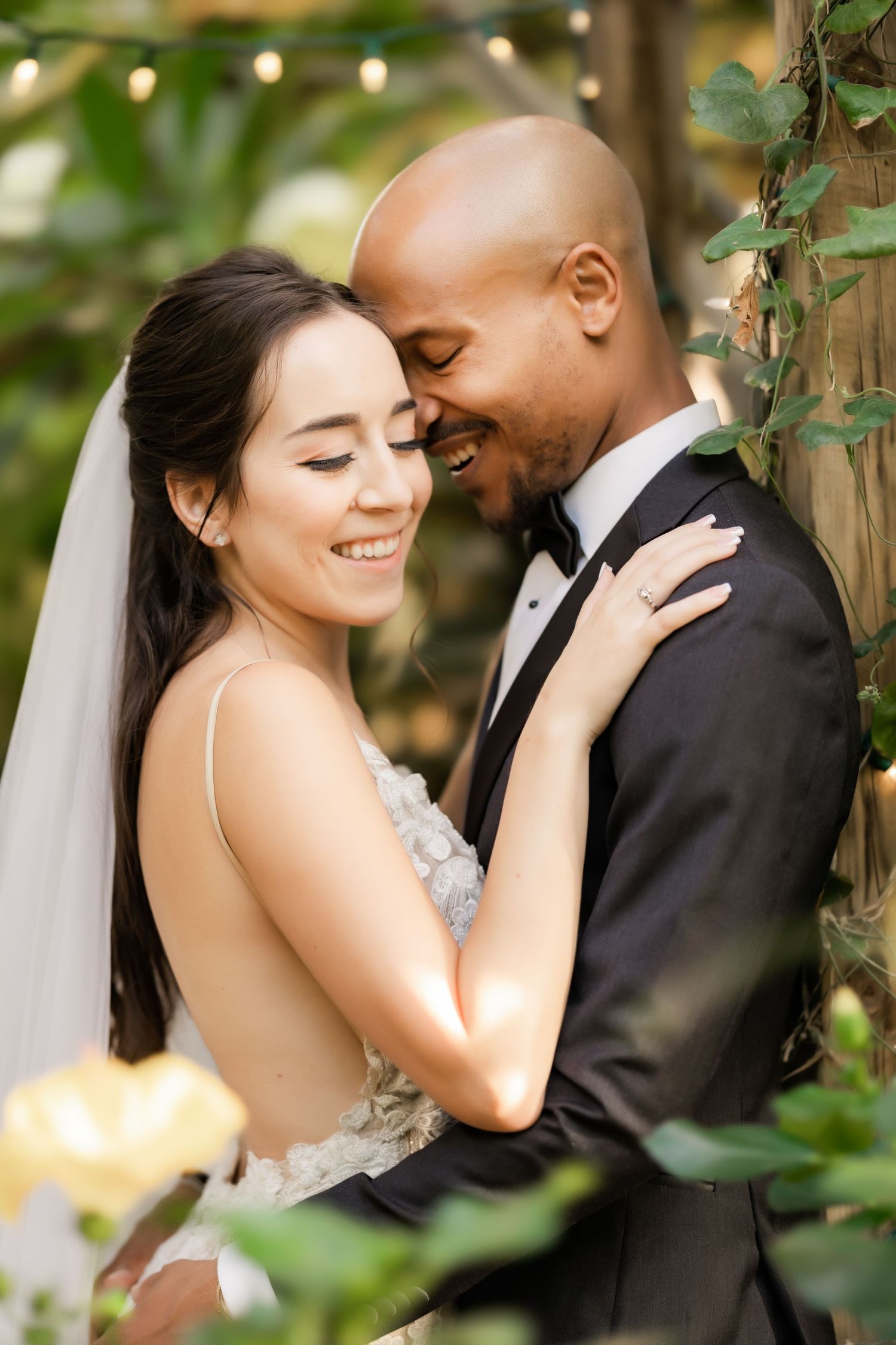 Newlywed couple shares an intimate moment in a garden setting with string lights and greenery.