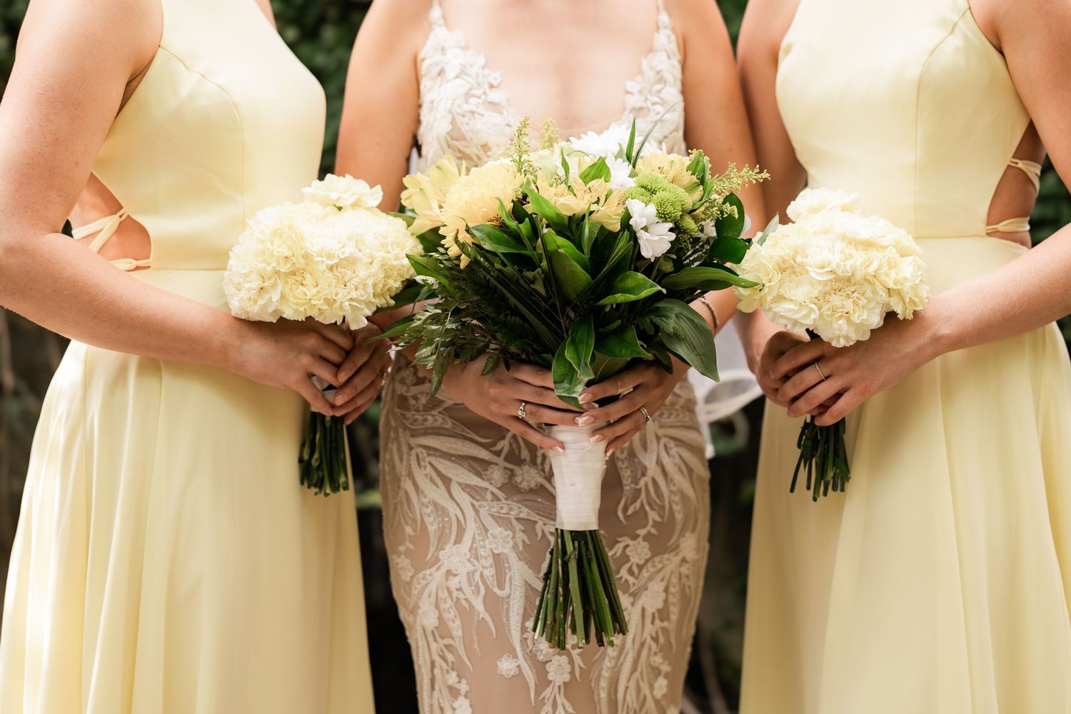 Bridesmaids in pale yellow dresses hold white flower bouquets next to bride in lace gown.