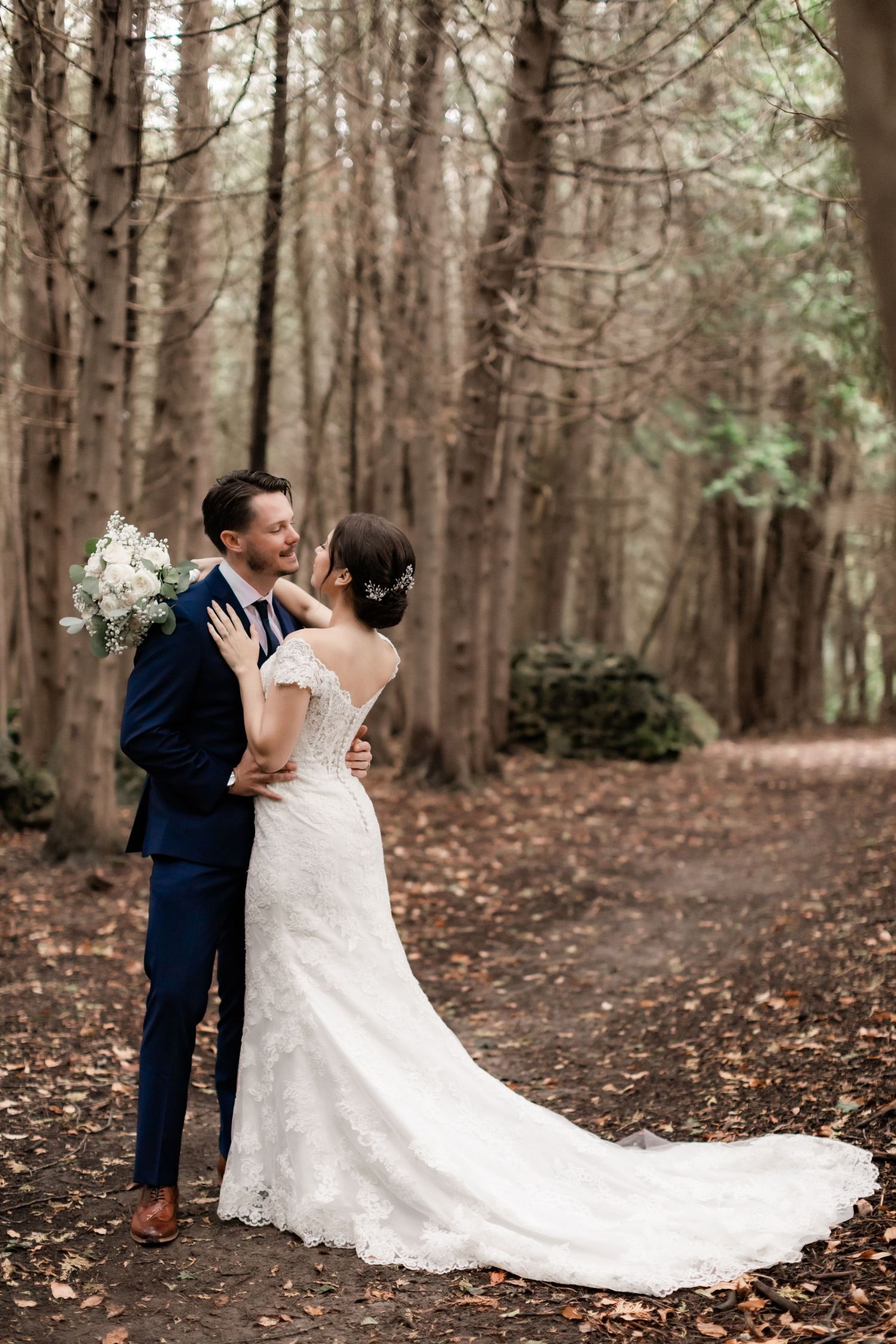 Wedding couple shares intimate moment on forest path with bride's lace train flowing behind.