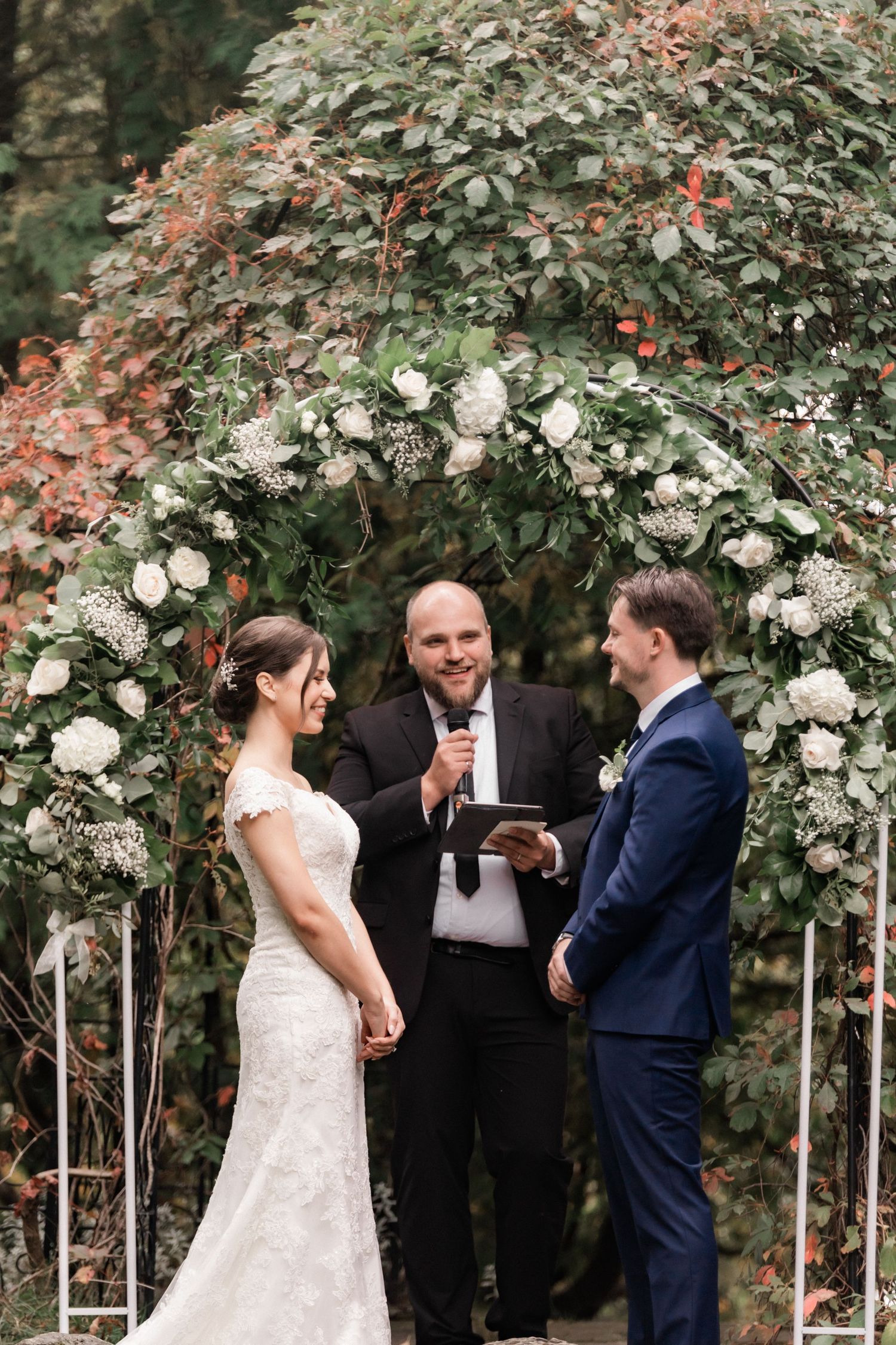 Wedding ceremony taking place under an elegant white floral arch in a garden setting.