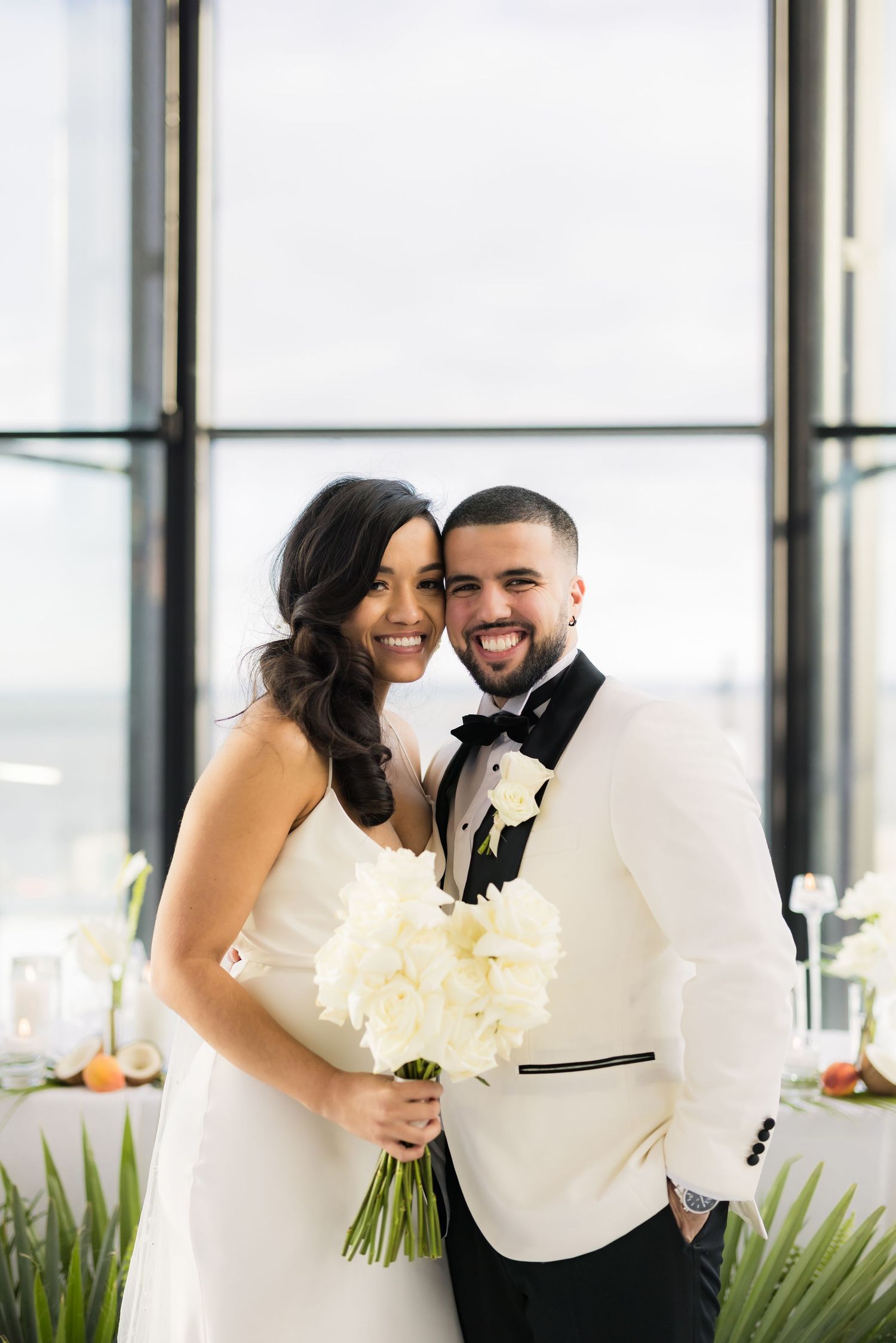 A happy couple shares a joyful moment in white wedding attire by large windows.