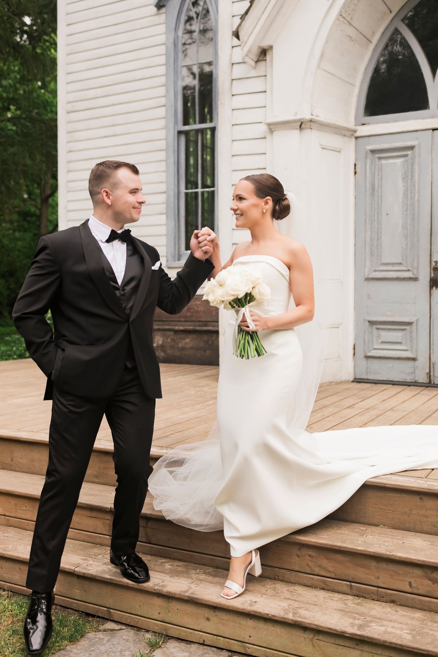 A playful wedding couple shares a joyful moment on wooden church steps outdoors.