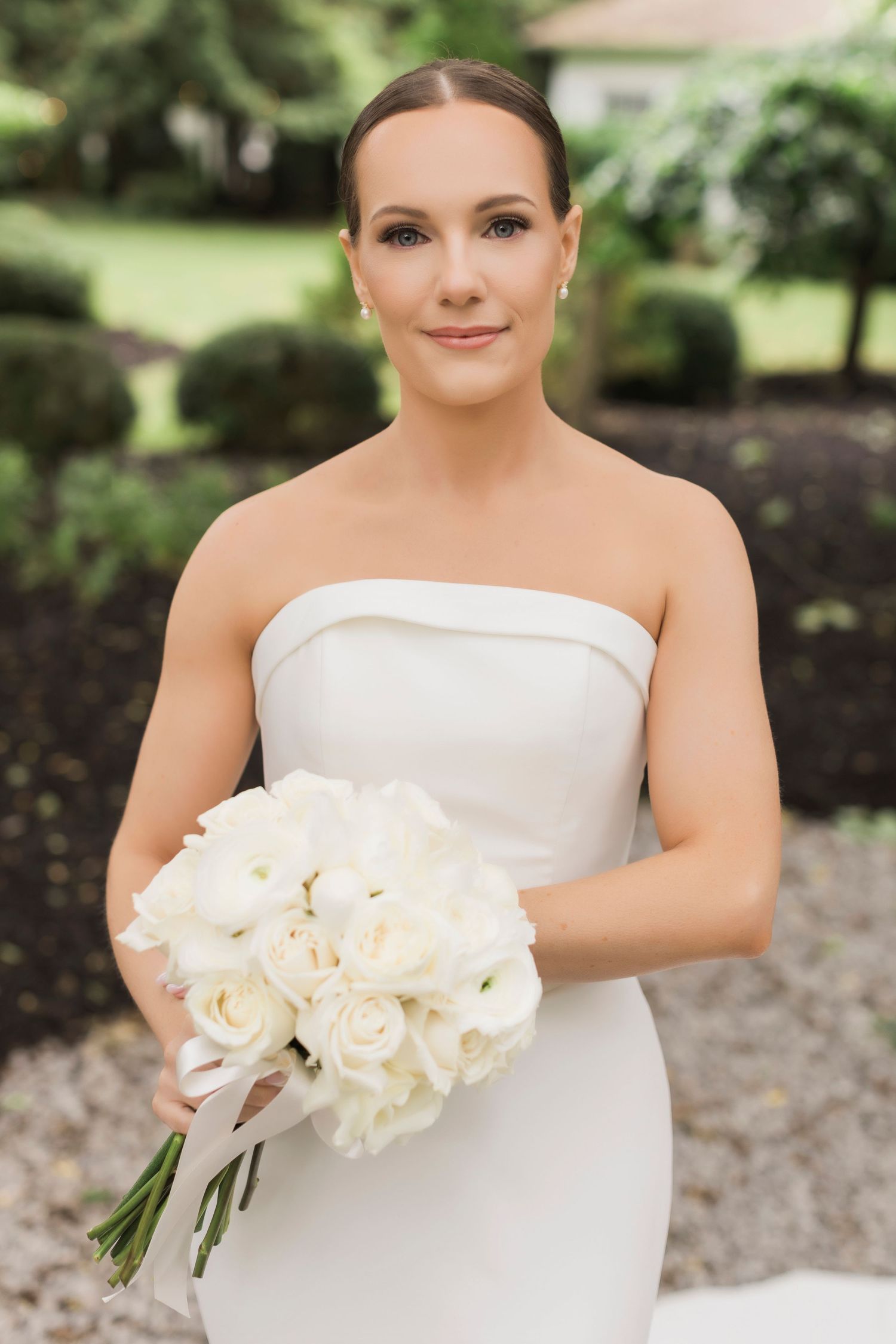 Elegant bride in strapless white gown holds classic white rose bouquet against garden landscaping.