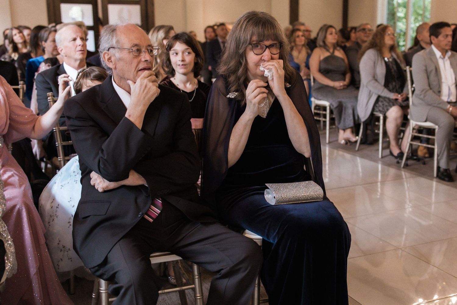 Wedding guests watching ceremony intently from white chairs in an elegant venue.