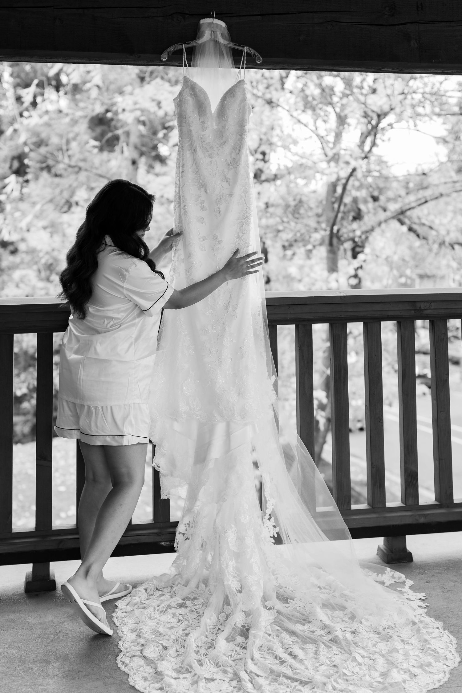 A silhouette examines a white wedding dress with detailed train hanging on a covered porch.