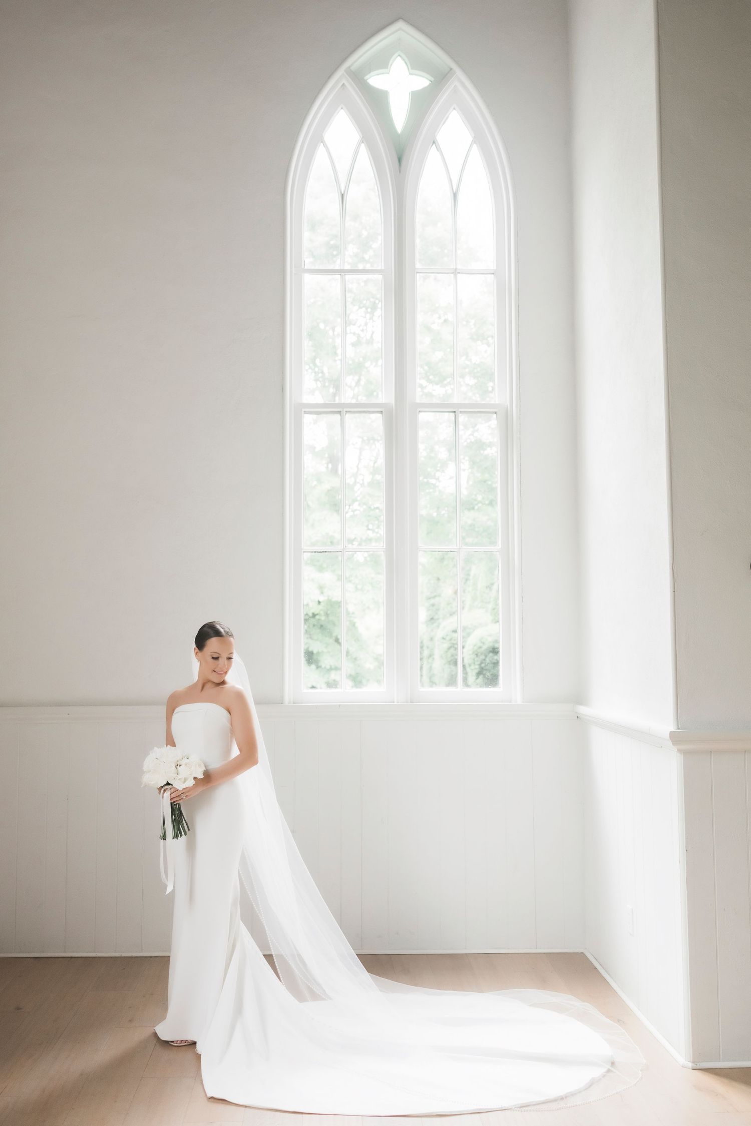 A white wedding gown with long train stands elegantly against a bright gothic window in a minimal white church interior.