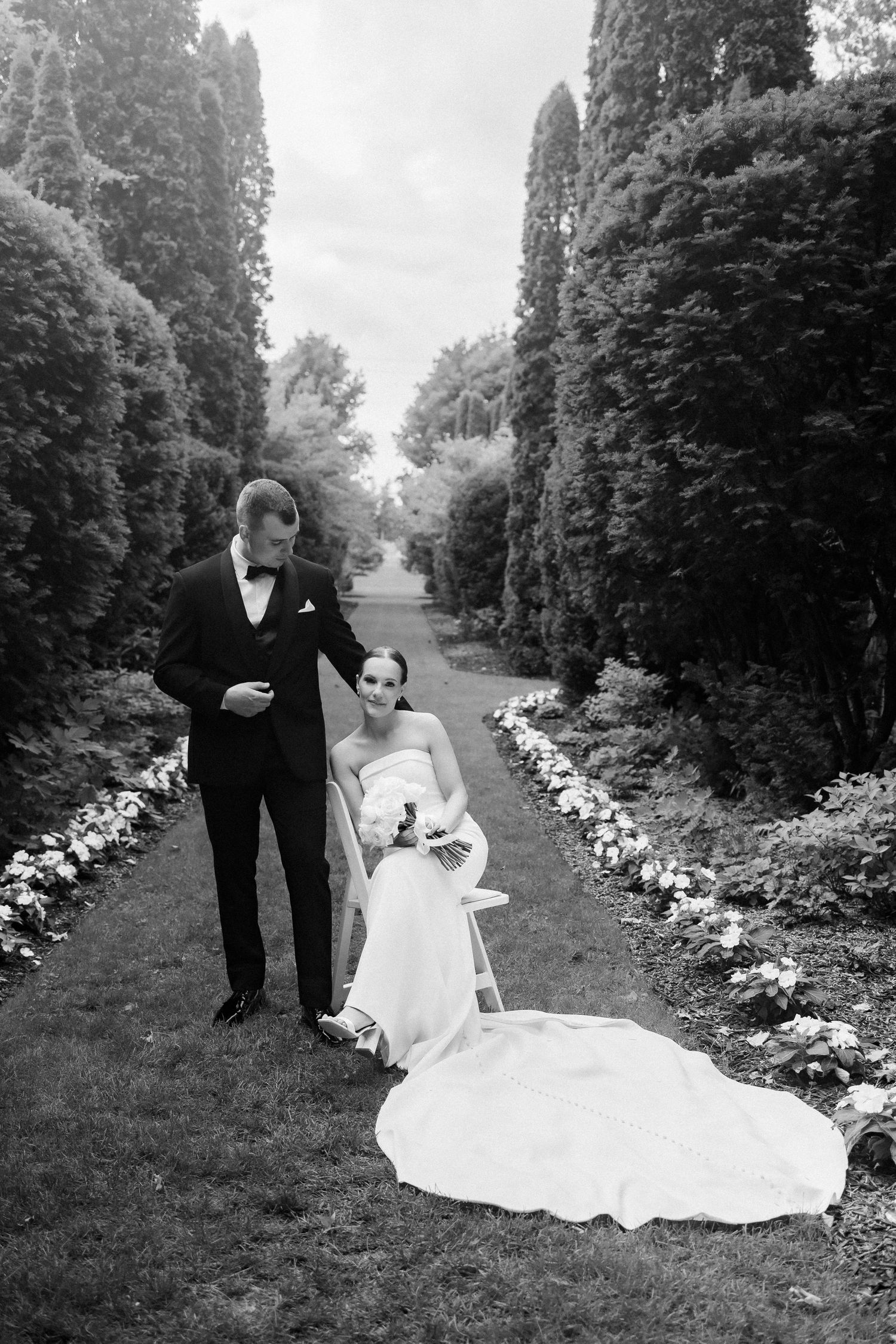 Black and white photo of newlyweds standing in garden path lined with tall cypress trees and flowers.