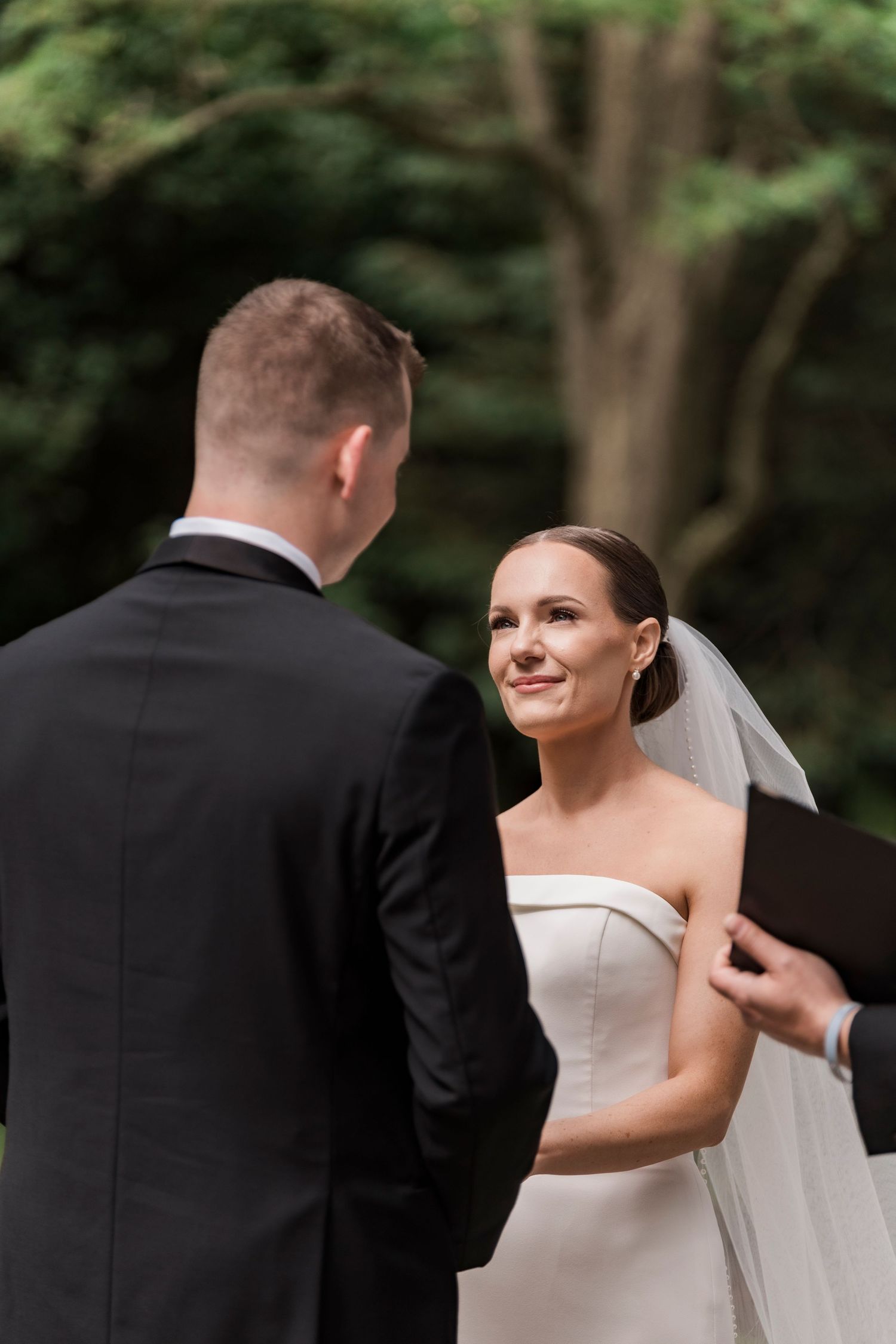 A bride wearing a strapless white gown and veil smiles during an outdoor wedding ceremony.