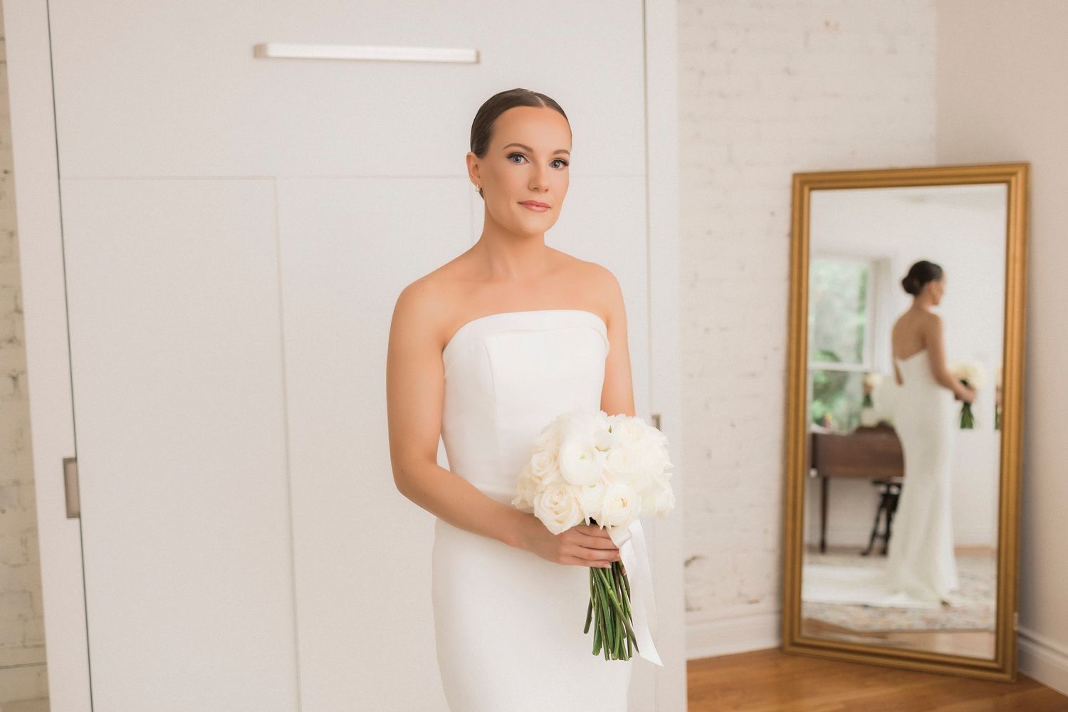 A bride in an elegant strapless white gown holds a classic white bouquet while standing in a bright room with a mirror.