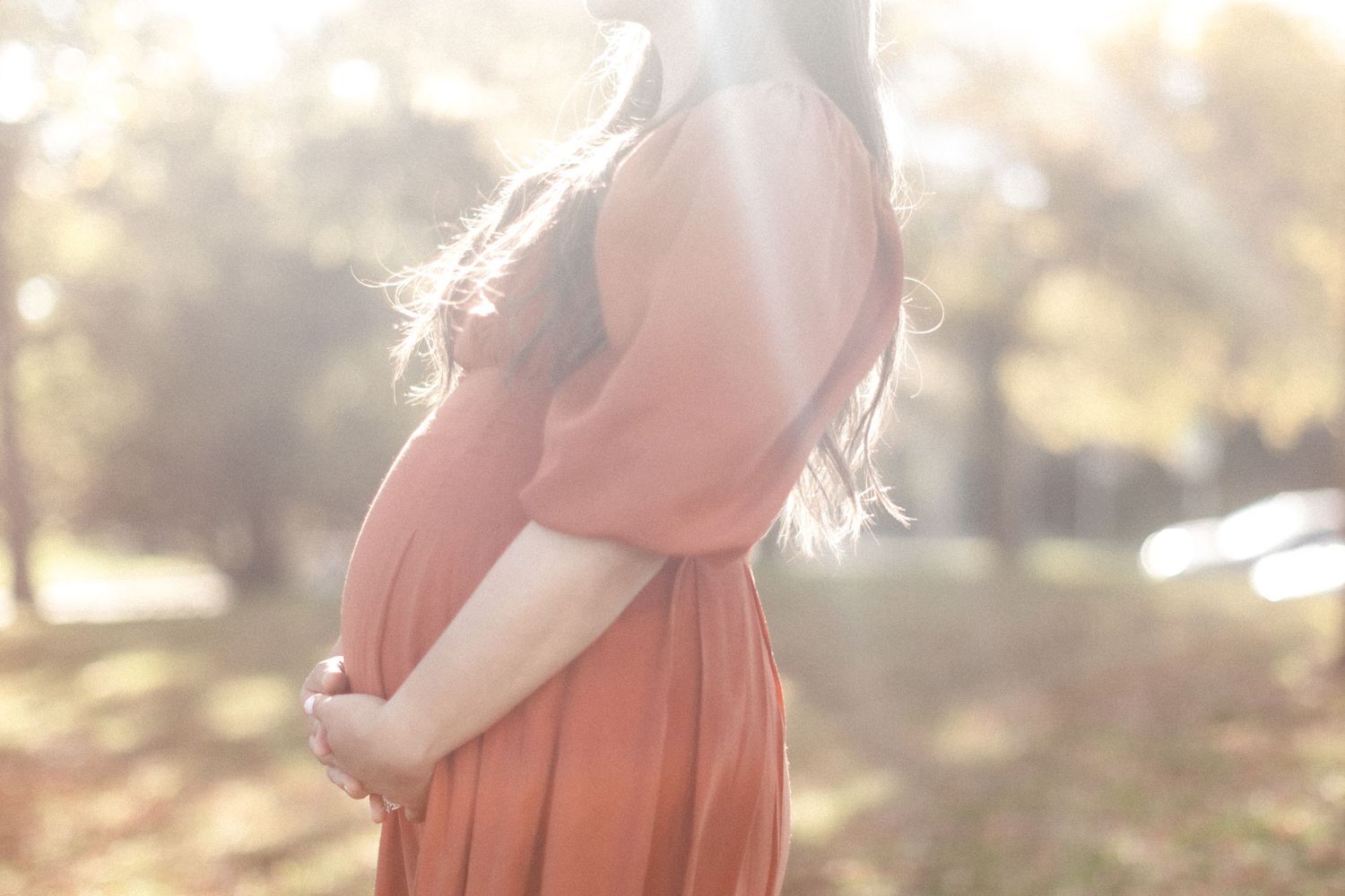 Silhouette of pregnant woman in flowing dress touching her belly in golden afternoon light.