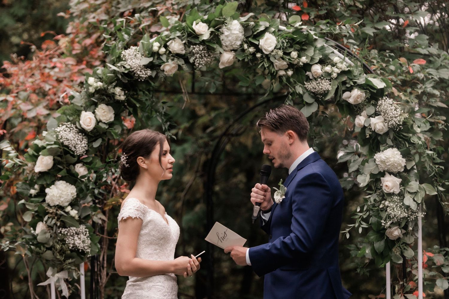 Wedding couple reads personal vows beneath elegant white rose archway in garden setting.