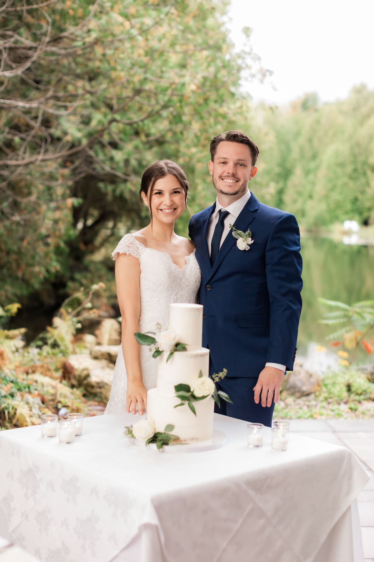 Newlyweds pose with their elegant white wedding cake adorned with fresh greenery outdoors.