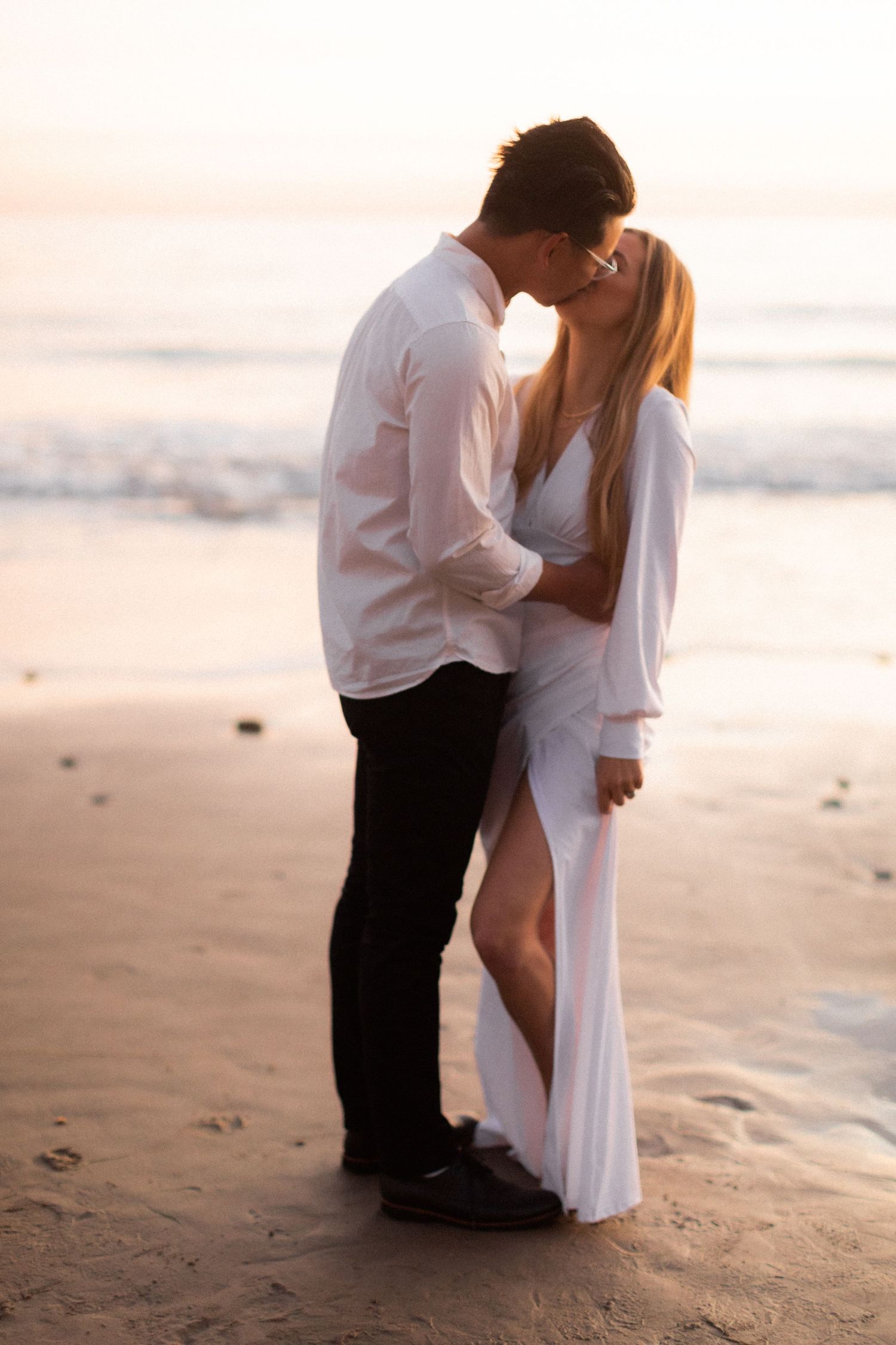 Couple sharing a romantic kiss on sandy beach at sunset with waves in background.