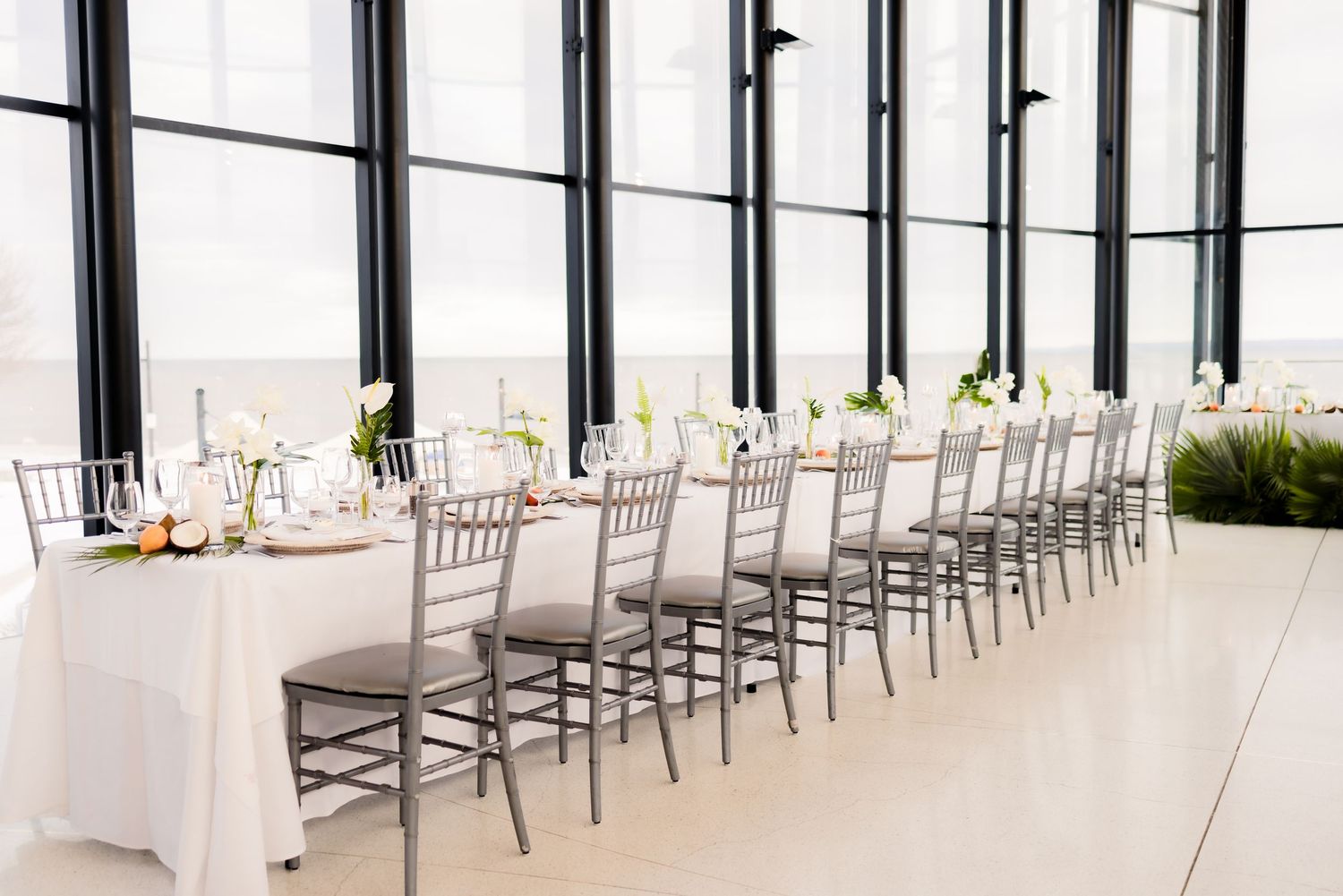 Long formal dining table with silver chiavari chairs and white linens set up in modern glass-walled venue.