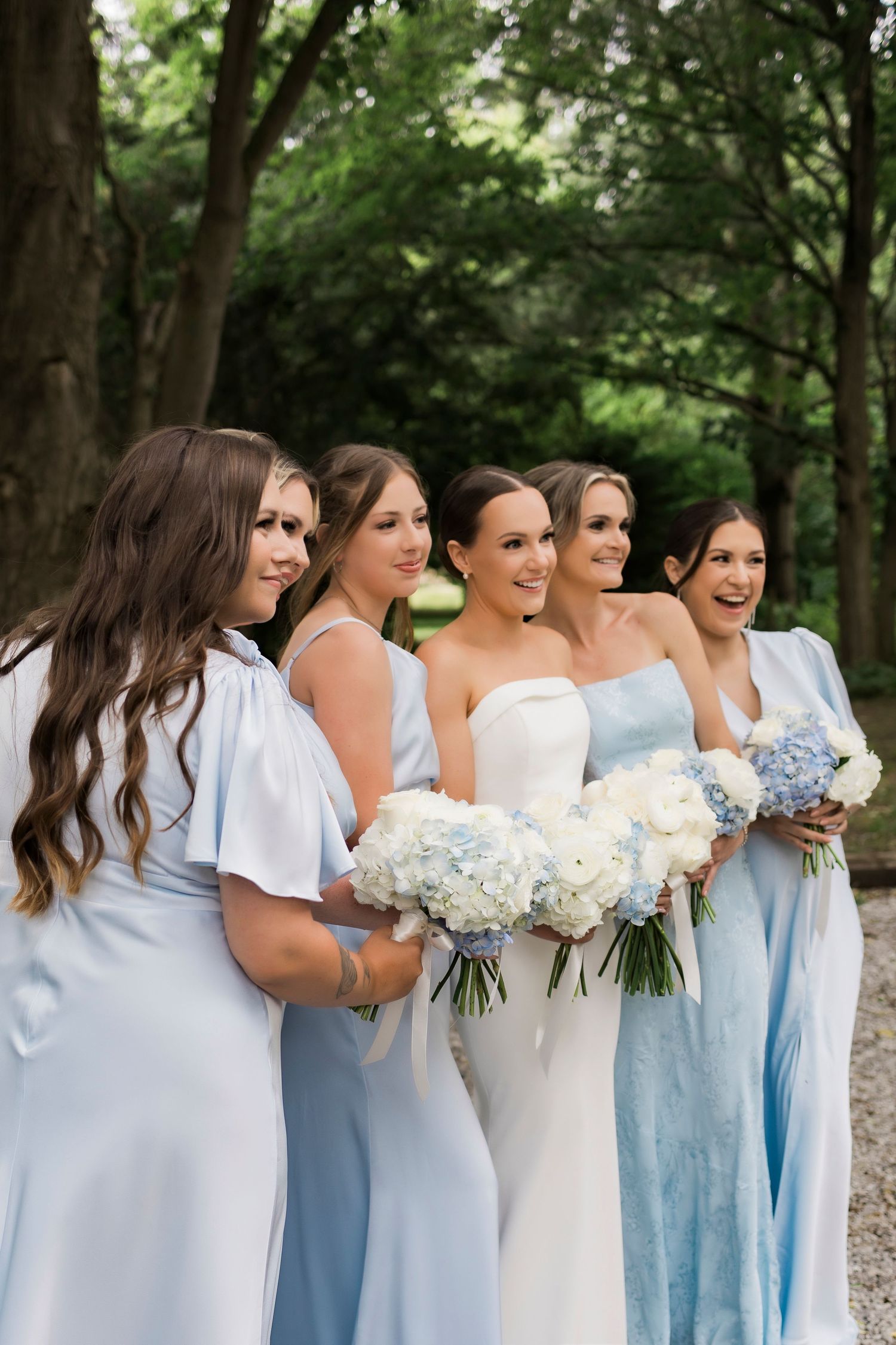 Wedding party in light blue bridesmaid dresses and white wedding gown hold hydrangea bouquets in outdoor garden setting.