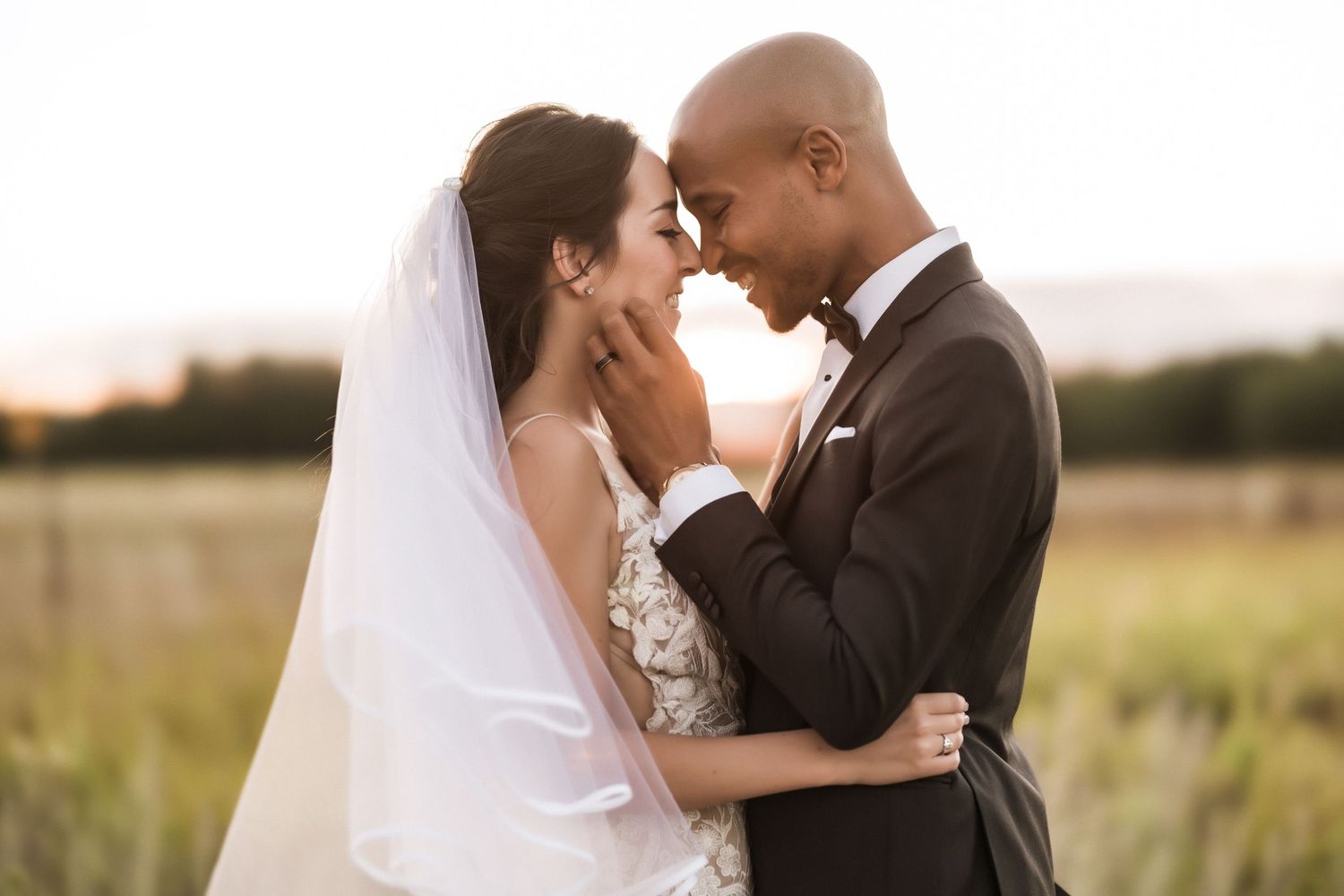 Bride and groom share an intimate embrace while standing in tall grass at sunset.