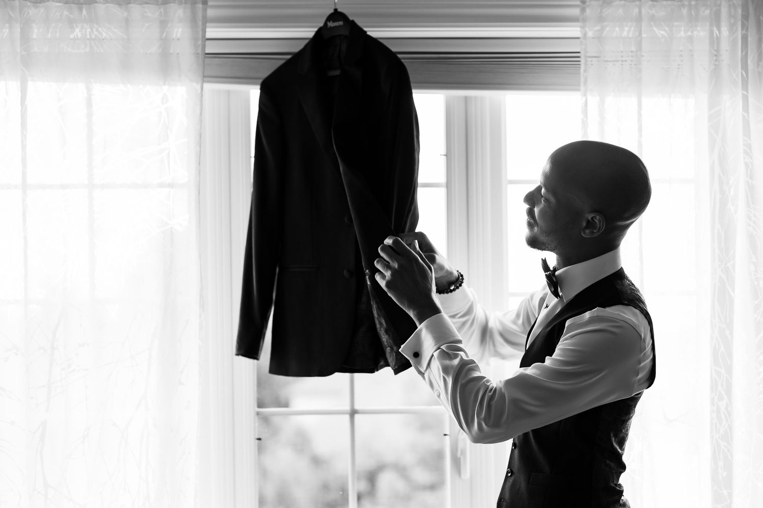 A black formal jacket hangs by a window in dramatic black and white lighting.