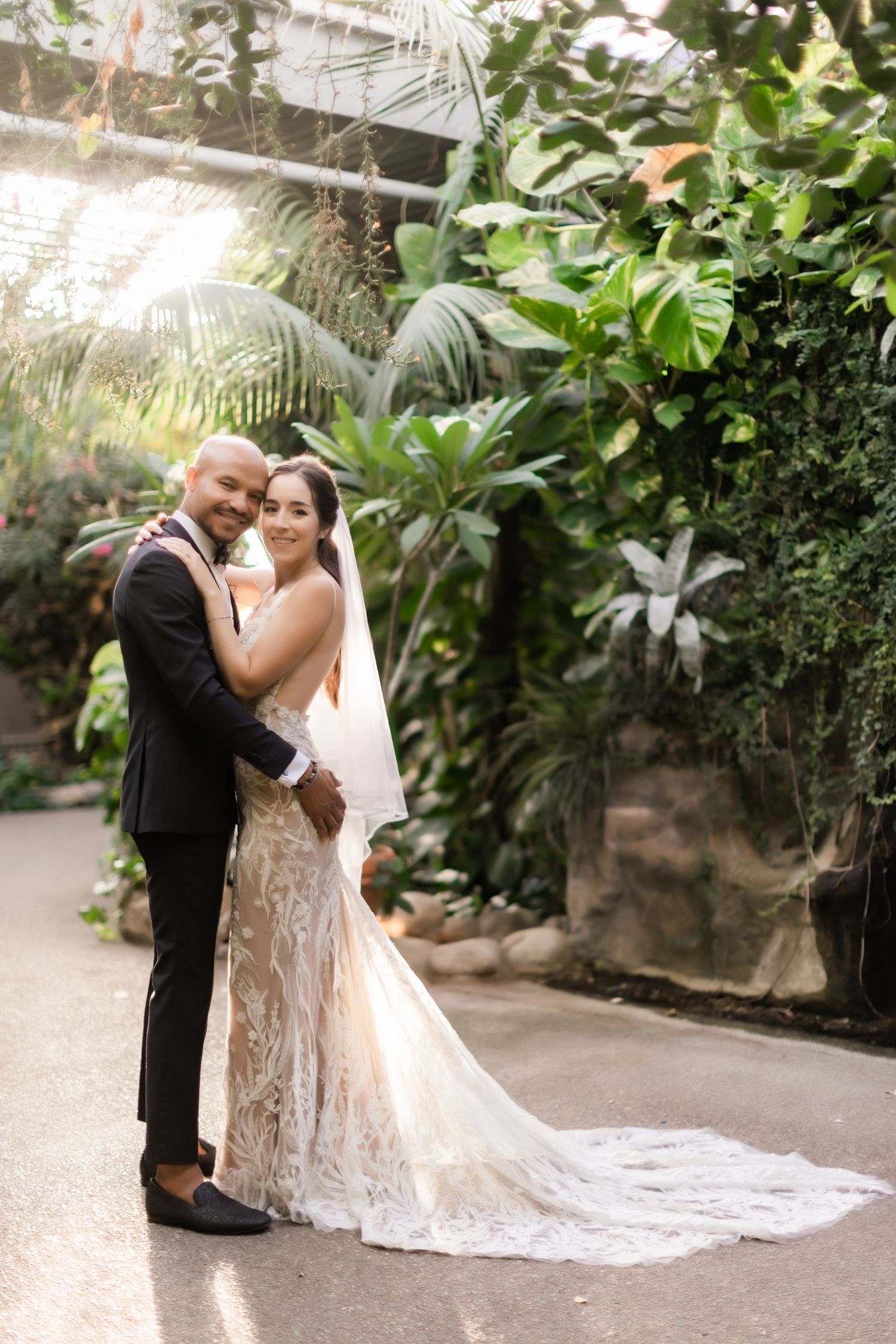 Wedding couple embraces on garden pathway with flowing lace dress train.