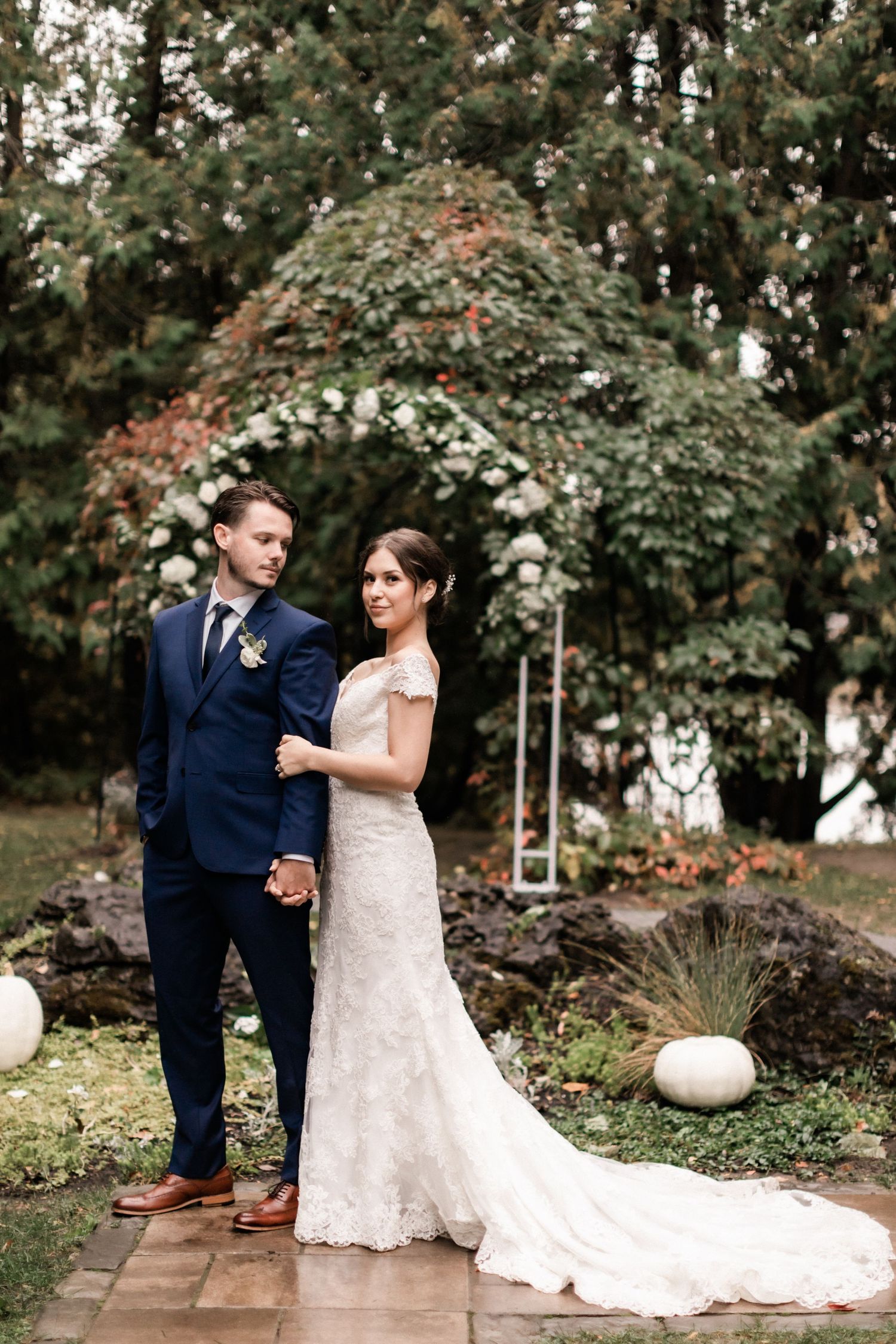 Wedding couple posing under garden arch with white pumpkins and brick pathway.