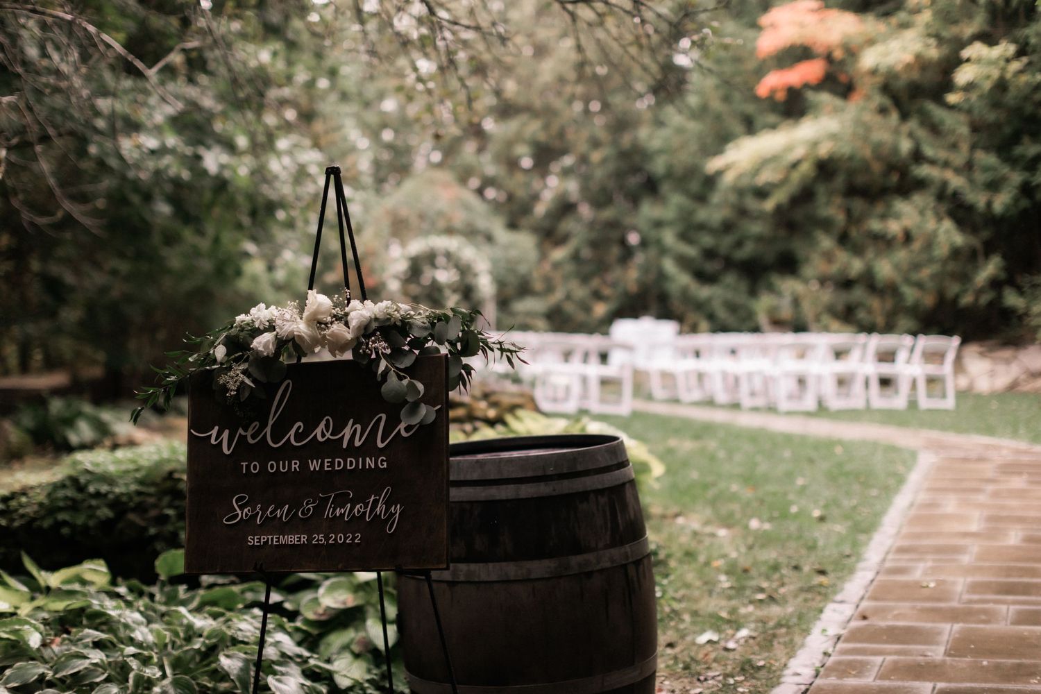 Rustic wedding welcome sign displayed on a wooden barrel with white chairs set up for an outdoor ceremony.