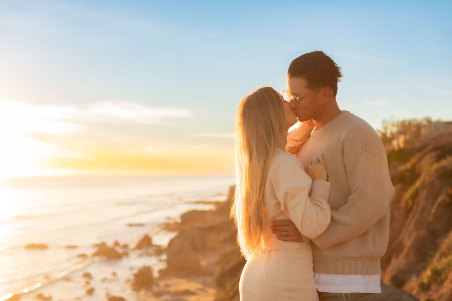 Couple embraces while watching sunset over rocky coastal cliffs on California beach.