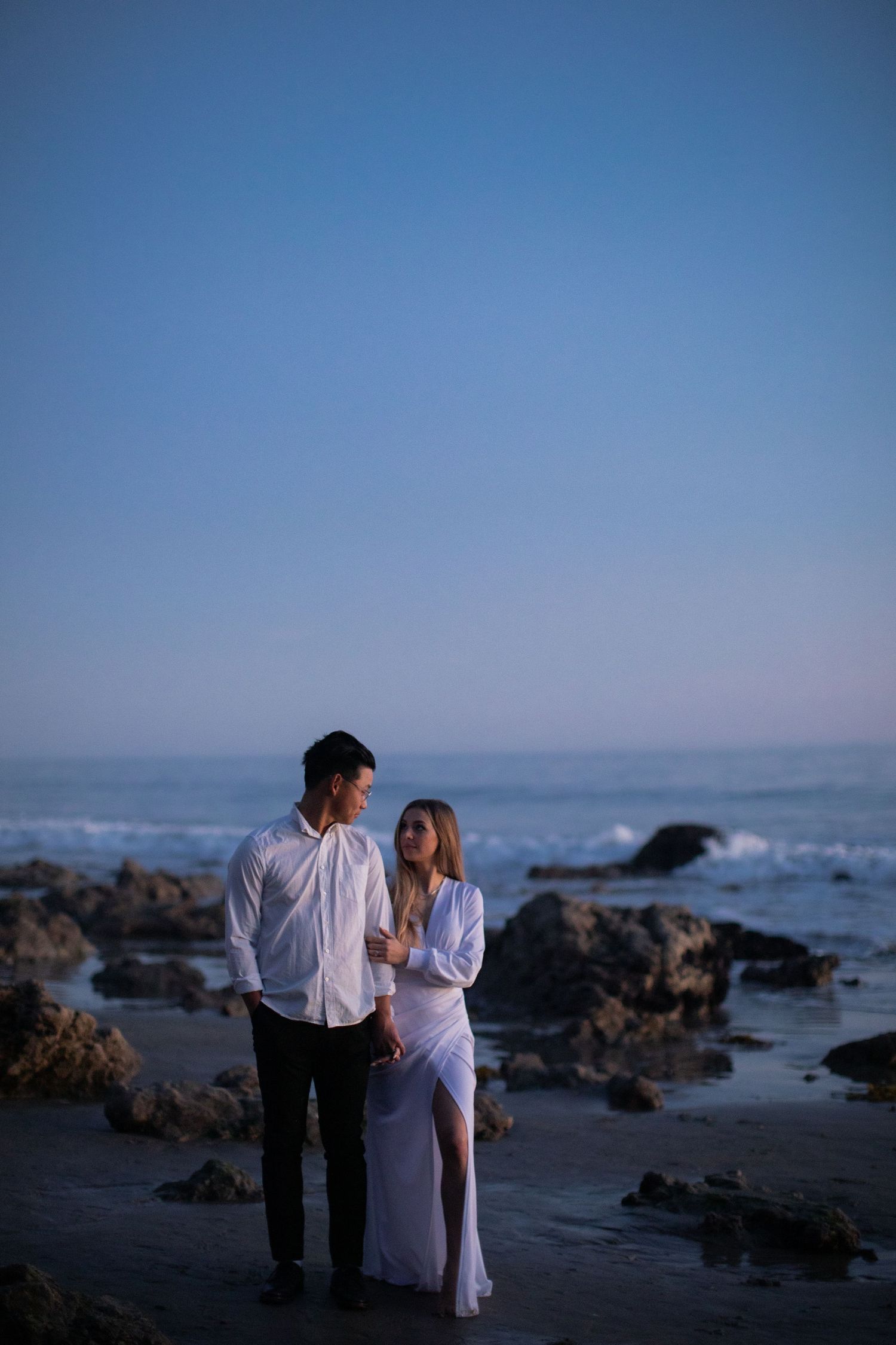 Couple in white attire sharing intimate moment on rocky beach at dusk.