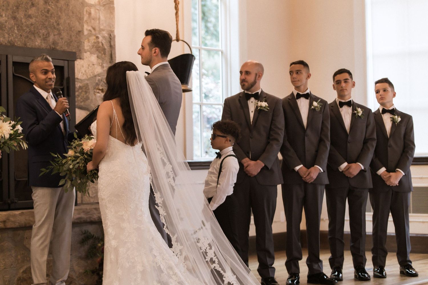 Wedding party lined up during ceremony with groomsmen in black suits and stone fireplace backdrop.