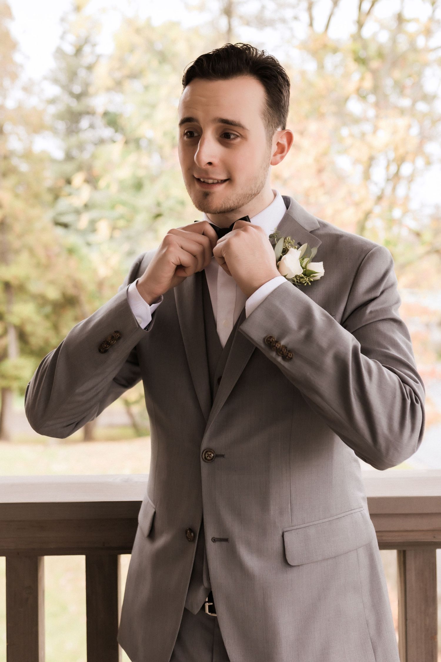 Groom adjusts his tie while wearing a gray three-piece suit with white boutonniere on a wooden porch.